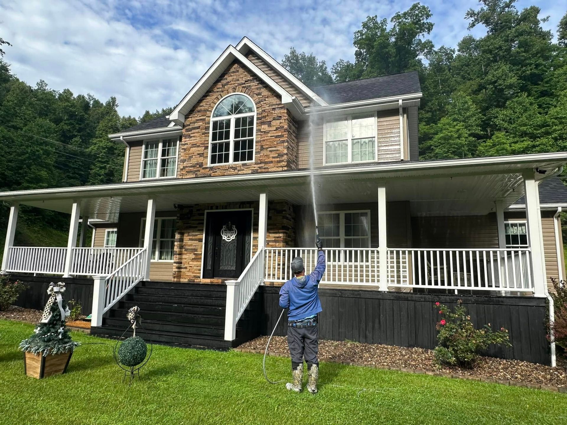A person stands on the lawn in front of a large stone house with a wraparound porch and dark shutters.