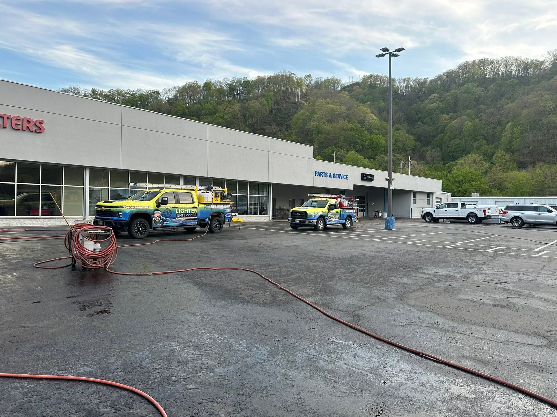 Shopping center parking lot with wet pavement and several utility trucks parked by a storefront.