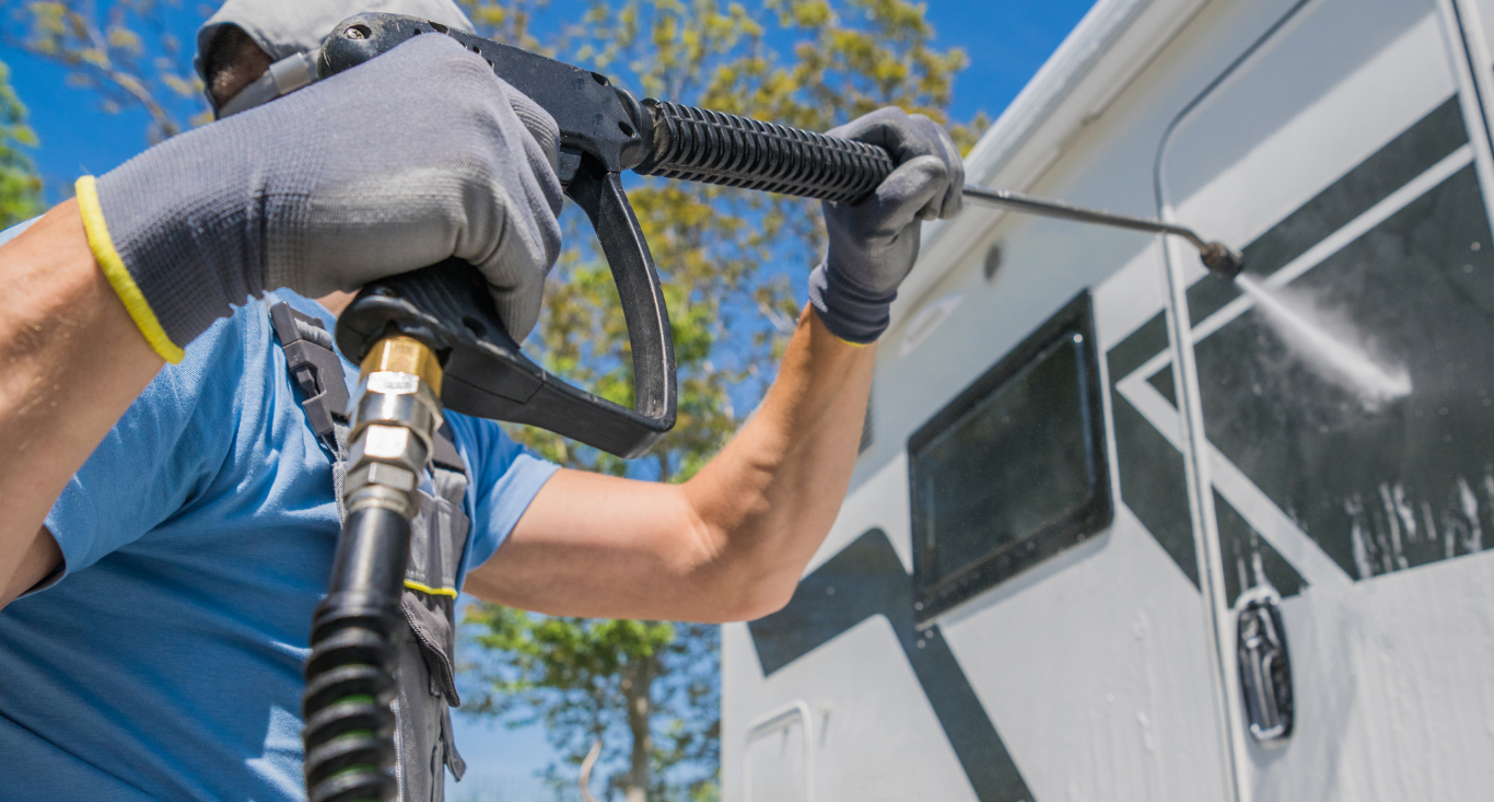 Gloved worker pressure-washing a white vehicle under a blue sky
