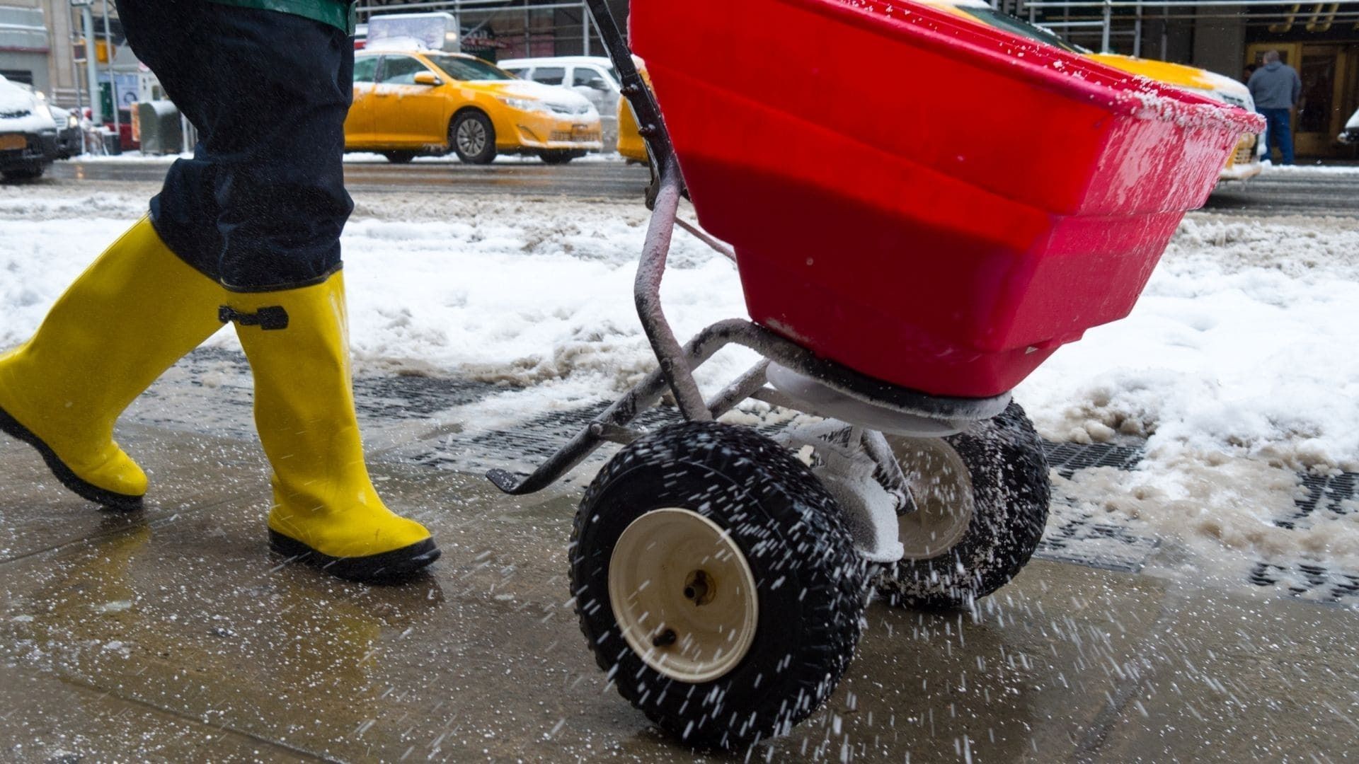 Person in yellow boots pushing a red cart through slushy snow on a city street