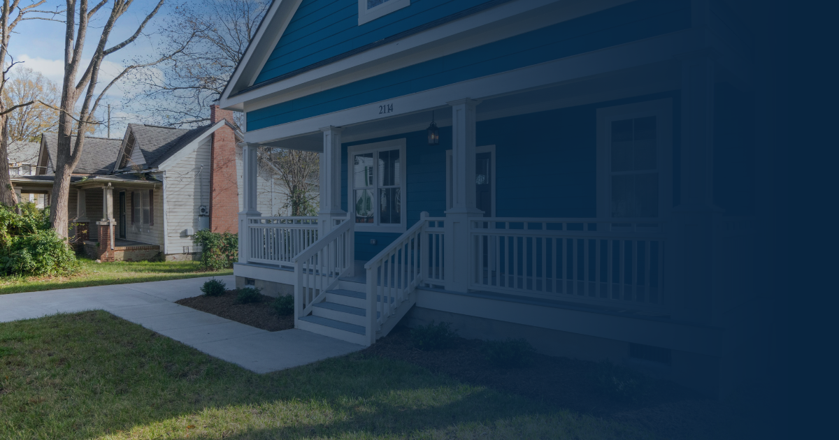 A blue house with a white front porch and railings, viewed from the front yard on a sunny day.