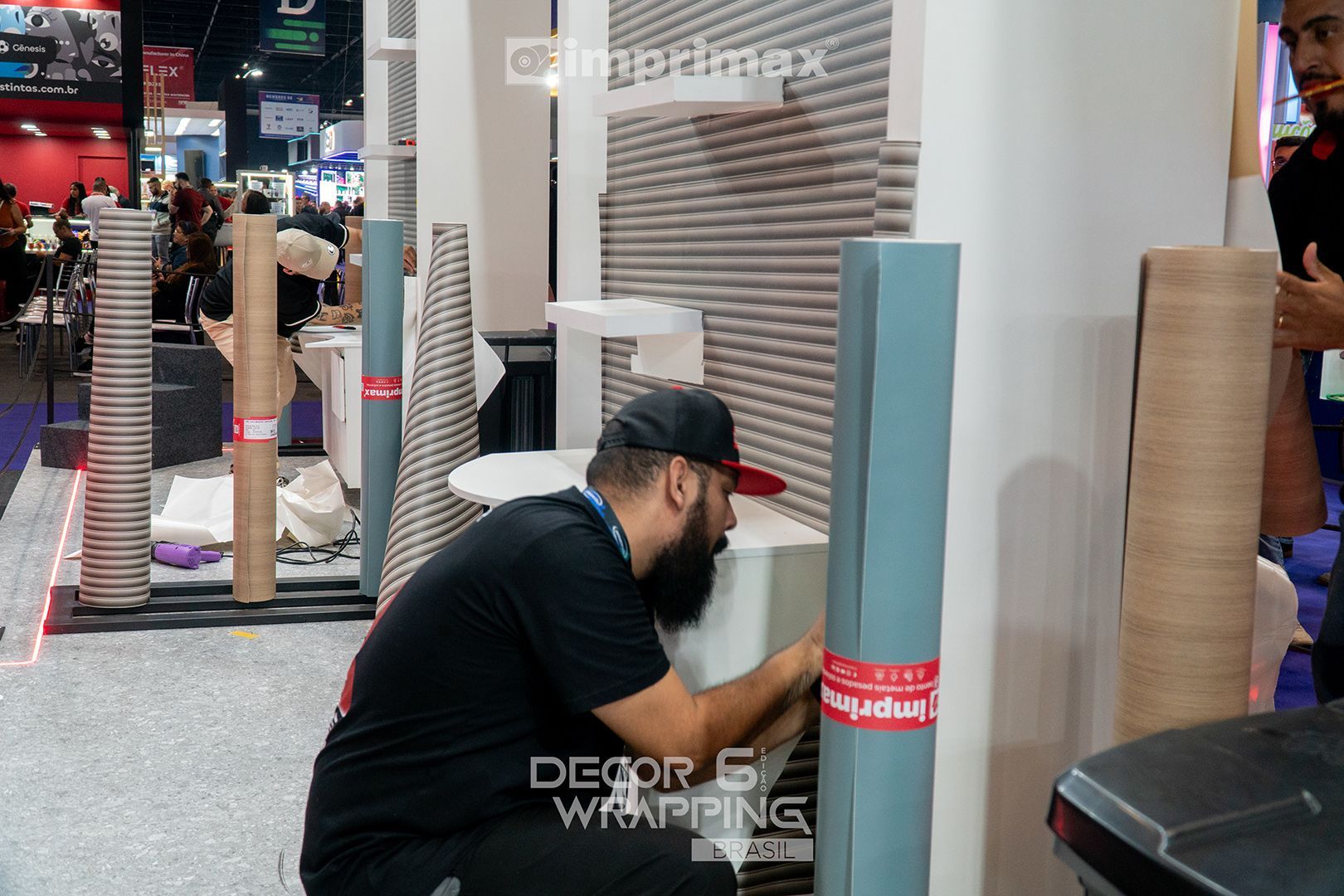 Um homem de camisa preta instala uma película adesiva em uma estrutura branca em um estande de feira comercial, onde rolos de material estão expostos.