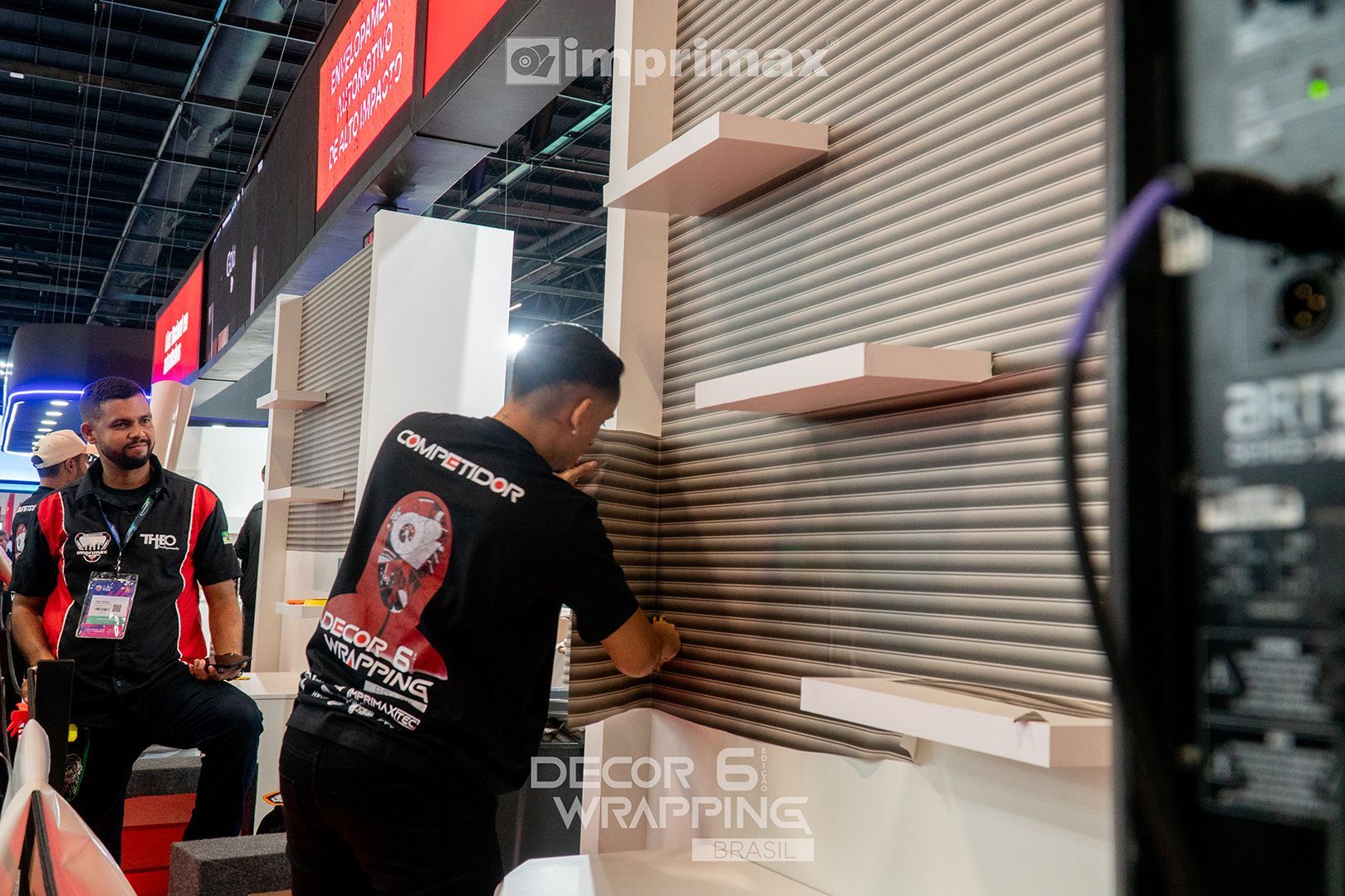 Um funcionário aplica um revestimento em uma parede texturizada com prateleiras embutidas em um estande da Imprimax Decor Wrapping em uma feira comercial.