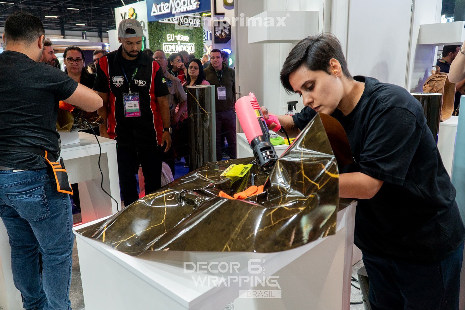 Uma pessoa usa um soprador térmico rosa para aplicar uma película adesiva escura com veios dourados em uma superfície em um estande de feira comercial interna.