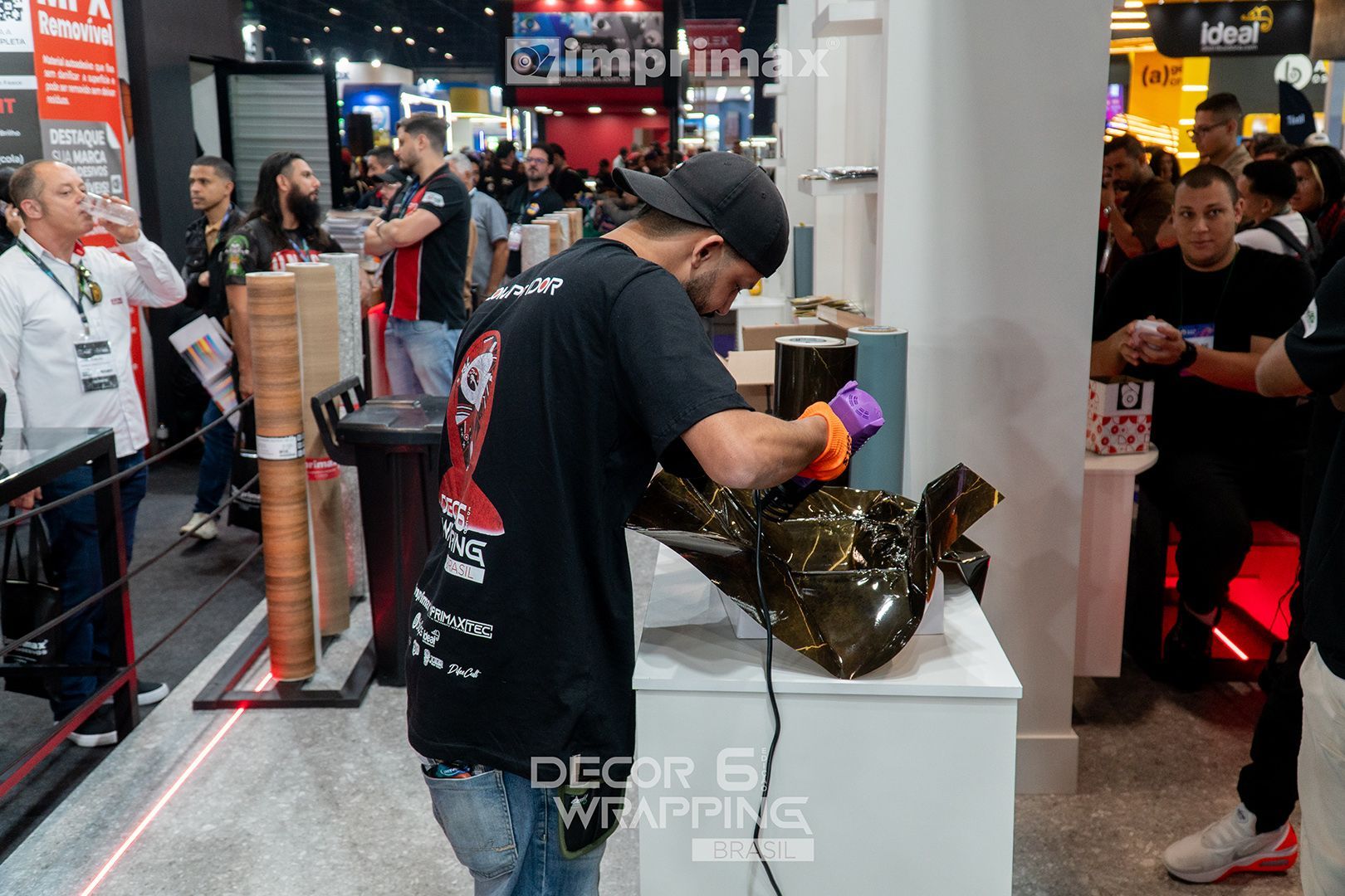 Uma pessoa de boné e camiseta pretos usa uma pistola de ar quente para aplicar vinil adesivo escuro em uma superfície curva em uma feira comercial lotada.