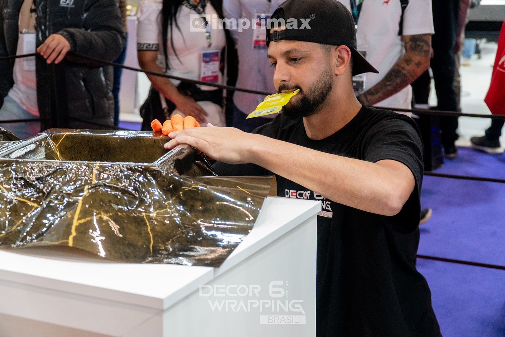 Uma pessoa usando boné e camiseta pretos aplica cuidadosamente uma película protetora dourada metálica em uma superfície branca em uma exposição.