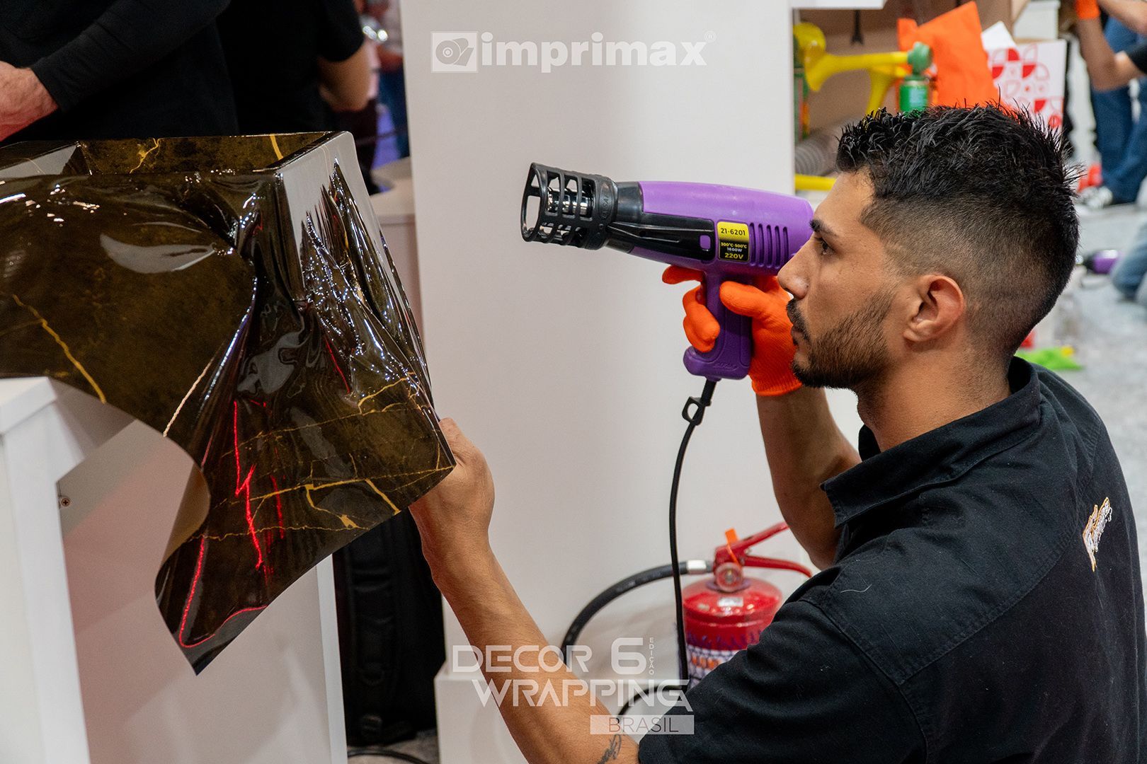Uma pessoa usando luvas laranja utiliza um soprador térmico roxo para aplicar uma película adesiva escura e refletora sobre uma superfície branca.