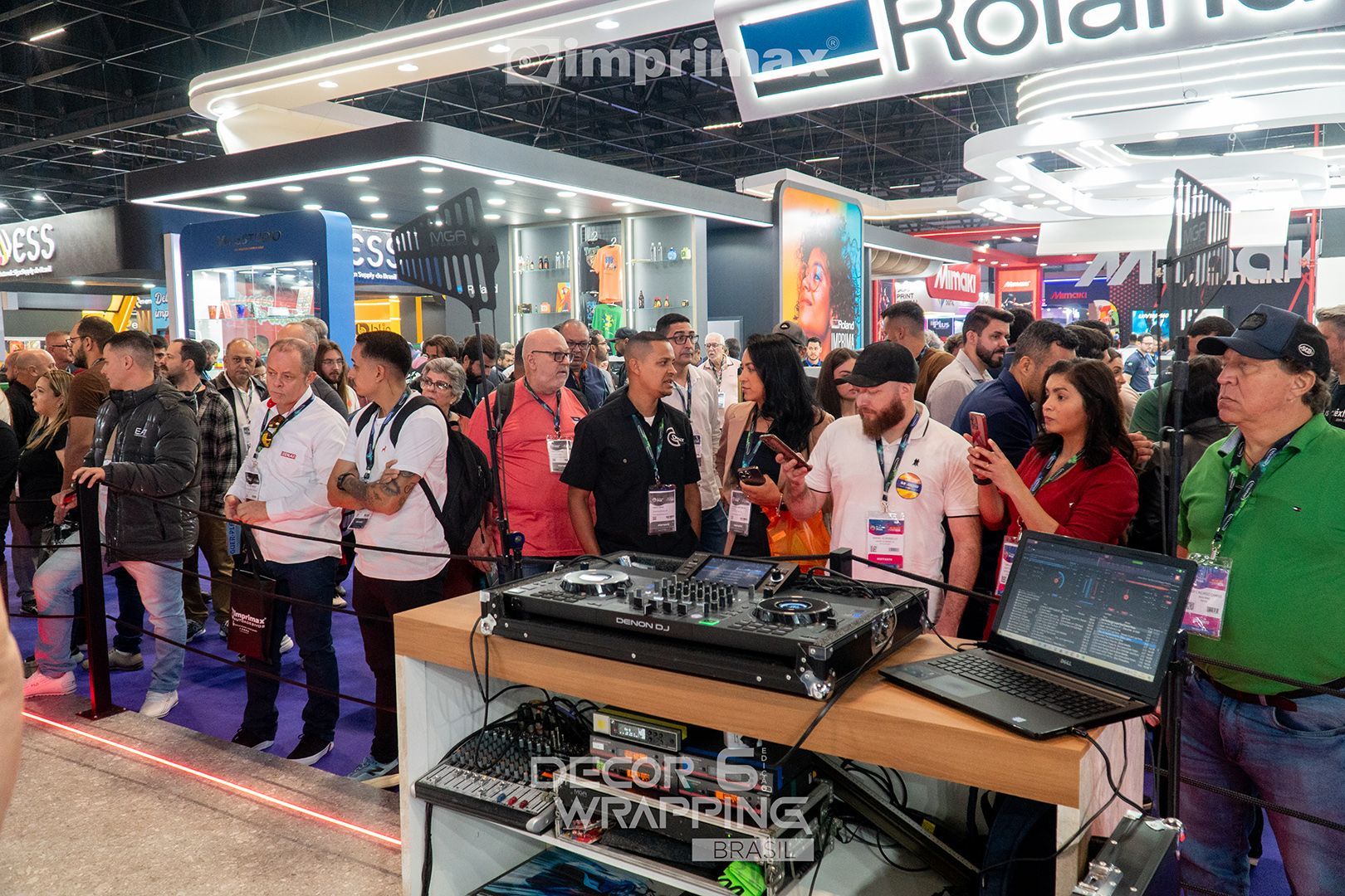 Uma multidão se reúne em volta de uma cabine de DJ com um laptop e uma mesa de som em um movimentado evento de feira comercial.