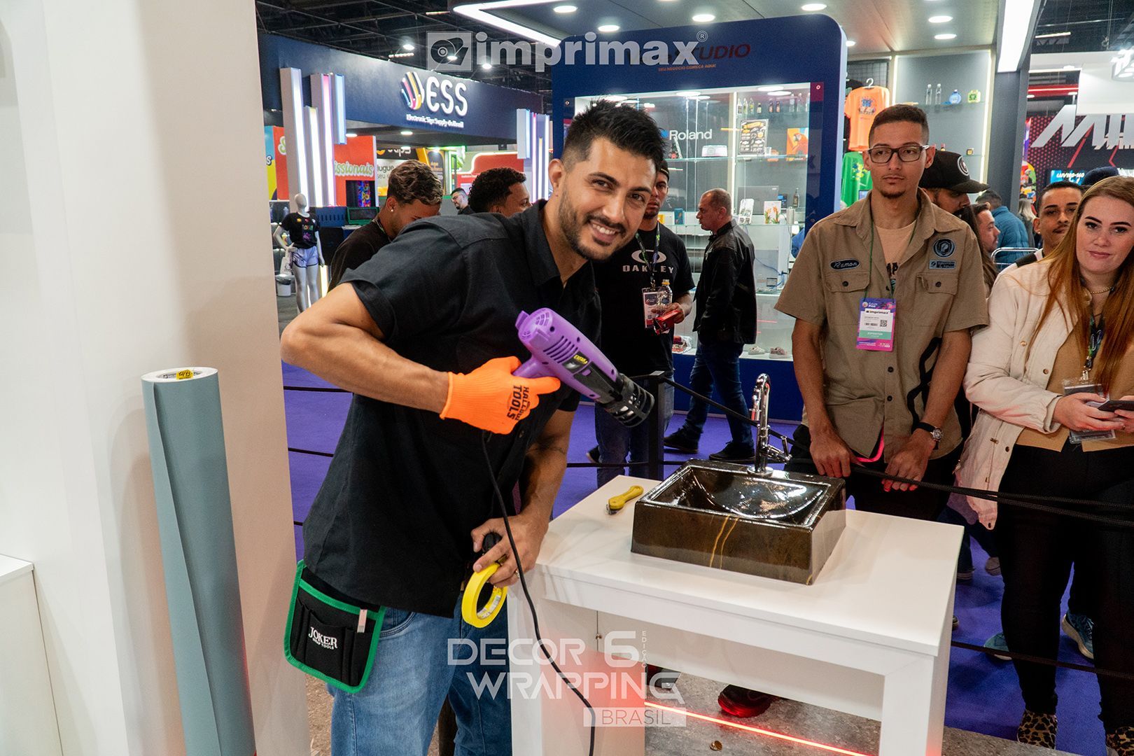 Uma pessoa usando luvas laranja utiliza um soprador térmico para aplicar um revestimento adesivo em uma mesa branca durante um evento de feira comercial.