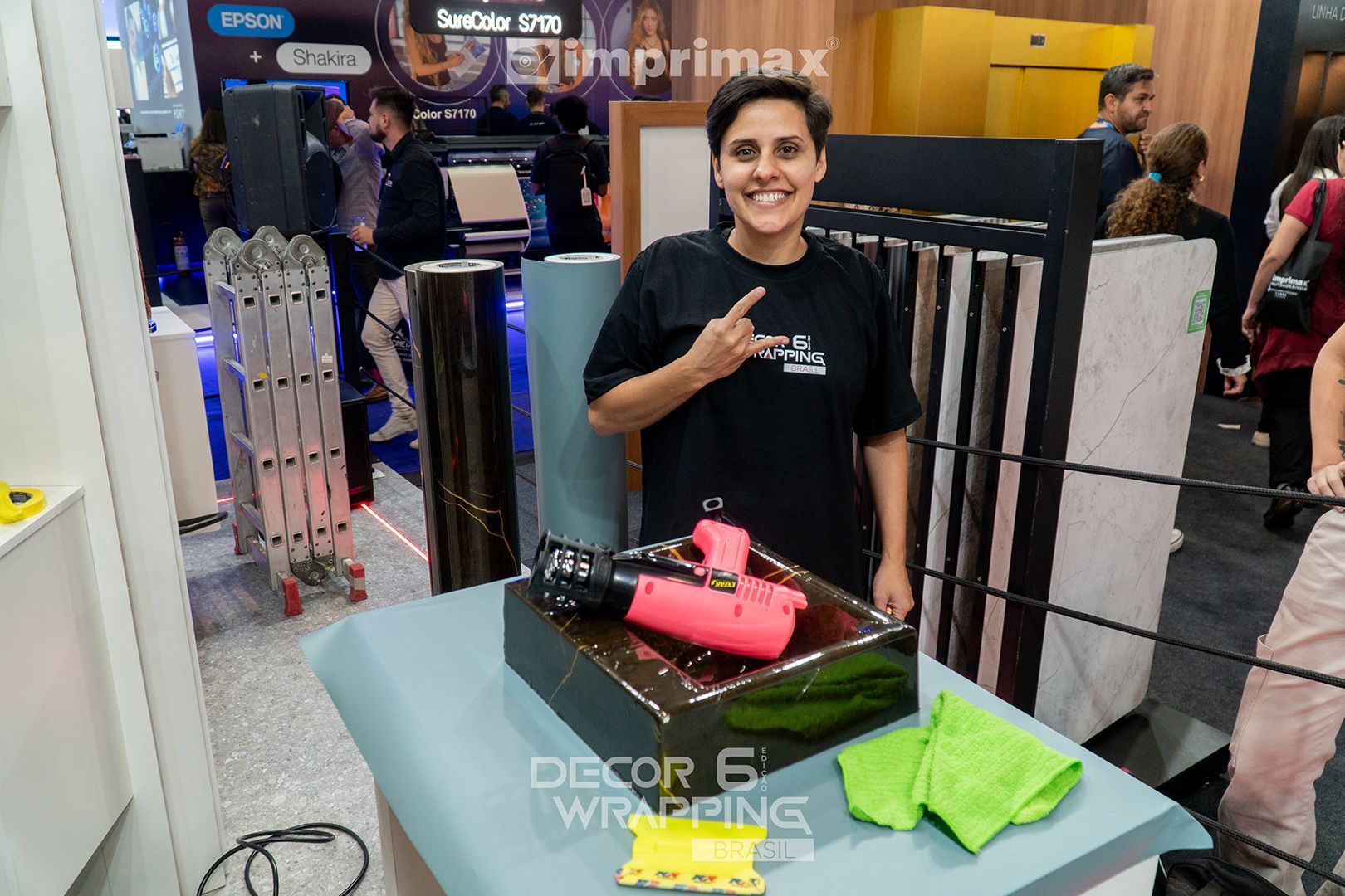 Um profissional sorridente em um estande de feira comercial, gesticulando perto de uma pistola de ar quente rosa apoiada em um bloco escuro envolto em plástico.