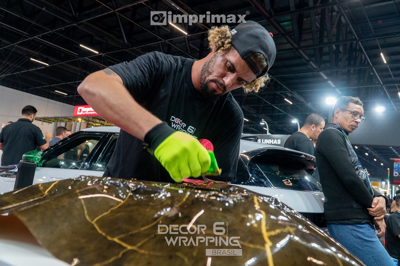 Uma pessoa vestindo camisa preta e luvas verde-claras aplica um adesivo vinílico marrom estampado em um carro em uma exposição em local fechado.
