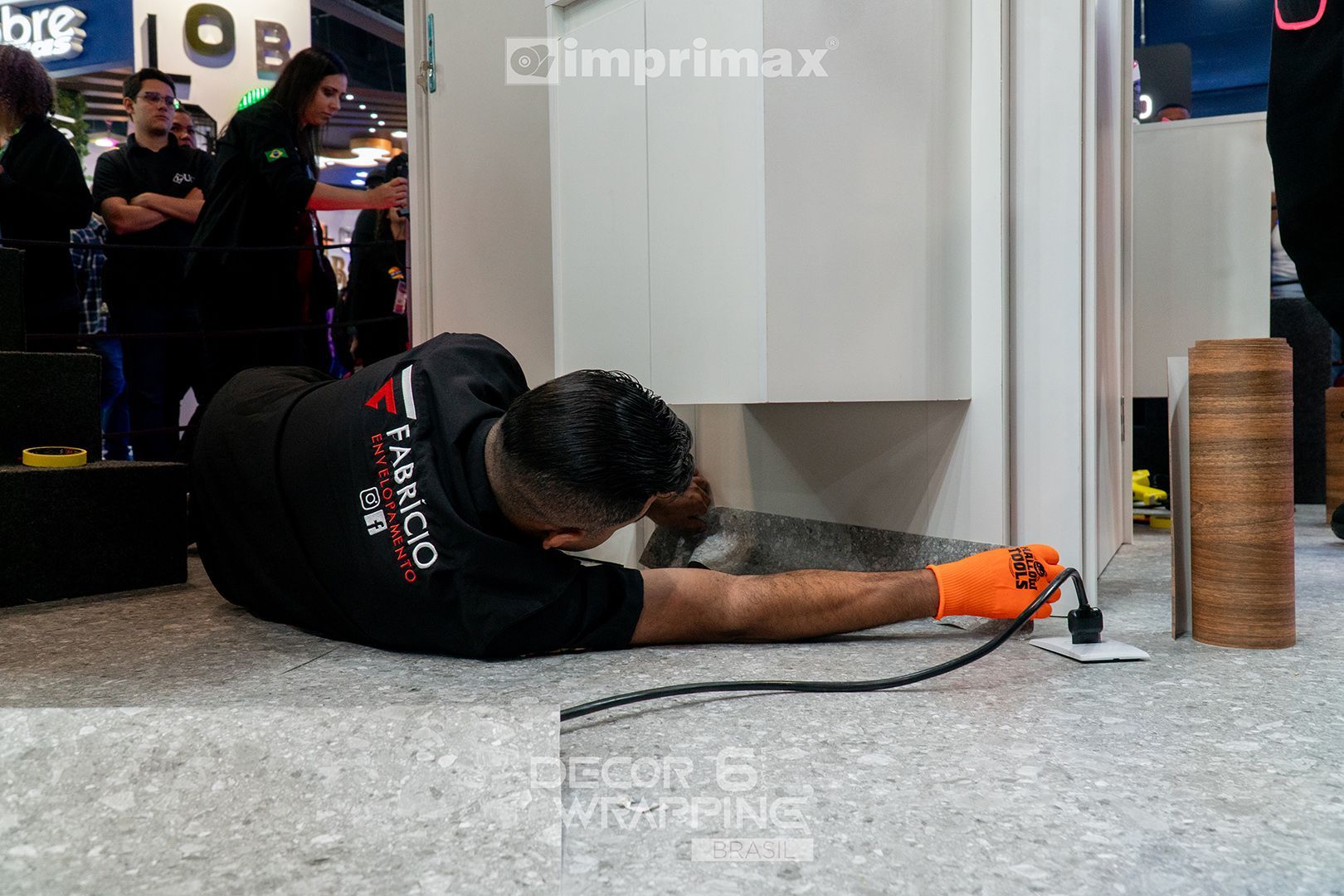 Um funcionário com camisa preta e luva laranja utiliza uma ferramenta de aquecimento no chão próximo a uma estrutura de exibição branca.