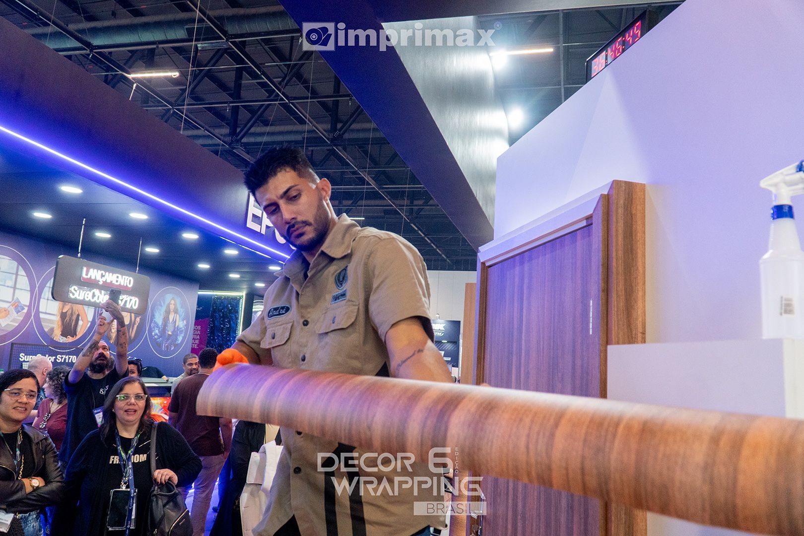 Um técnico com uniforme bege aplica um revestimento vinílico com textura de madeira em uma superfície no estande da Imprimax em uma feira comercial.