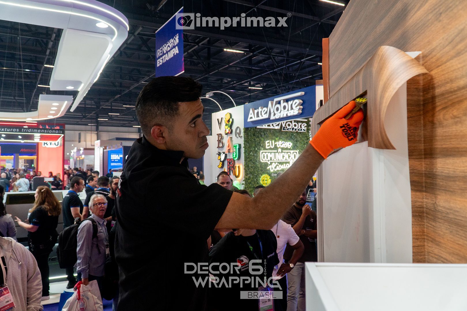 Um profissional aplicando película adesiva com textura de madeira em uma superfície em um estande de feira comercial, usando uma luva de segurança laranja.