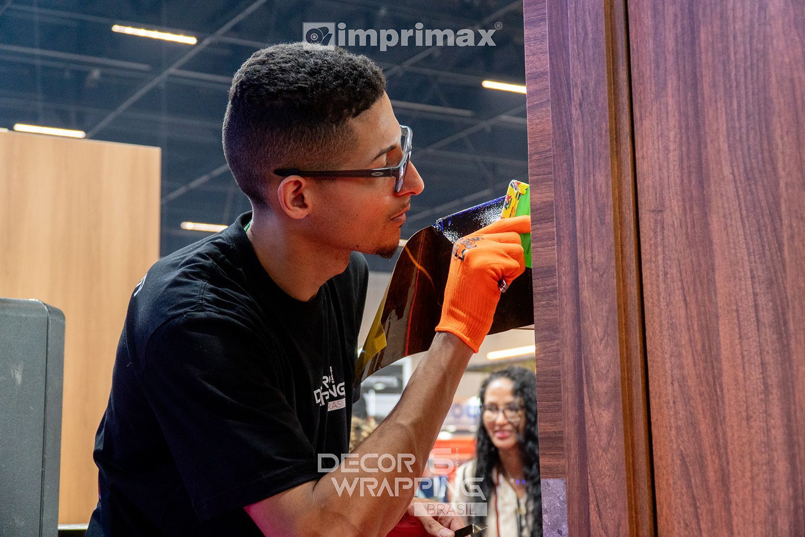 Uma pessoa usando uma luva laranja aplica cuidadosamente um revestimento vinílico com textura de madeira na superfície de uma porta em uma feira comercial fechada.