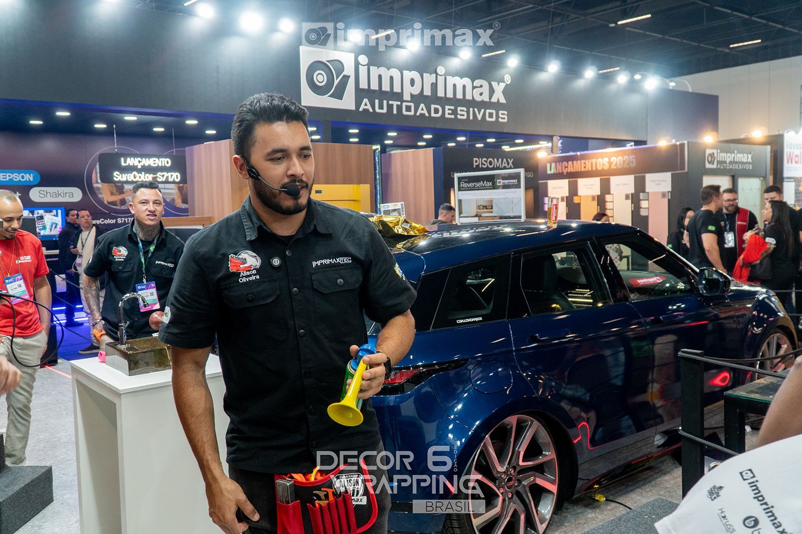Um apresentador segura uma pequena ferramenta amarela em um estande da Imprimax em uma feira comercial, em frente a um carro envelopado de azul.