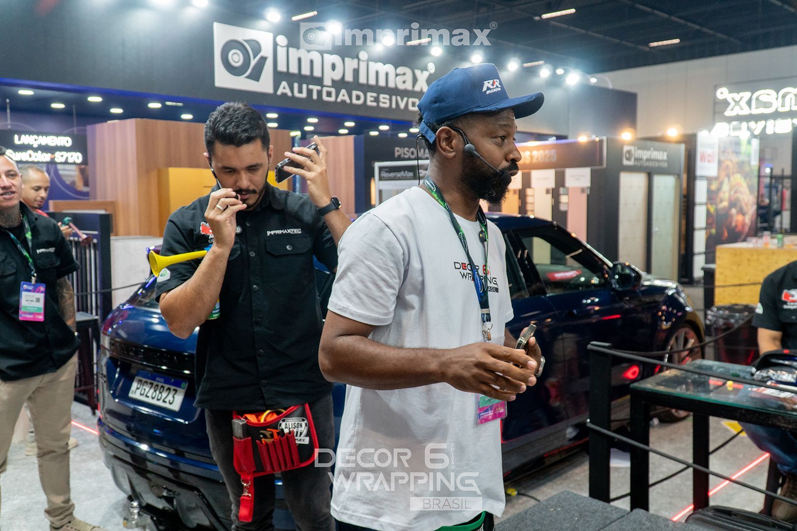 Duas pessoas trabalhando em um estande da Imprimax em uma feira comercial, uma segurando uma ferramenta perto de um carro azul, a outra olhando para o lado.