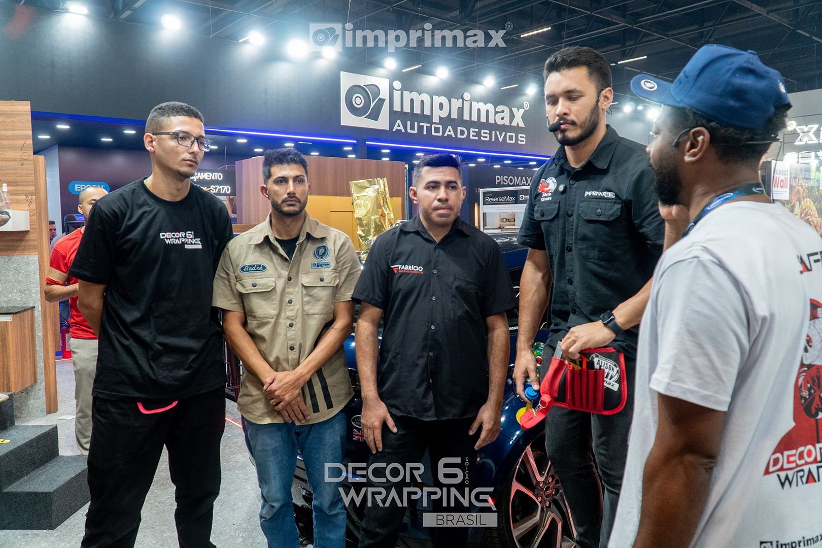 Grupo de pessoas em um estande da Imprimax em uma feira comercial, observando uma pessoa trabalhando em um projeto de envelopamento de carro.