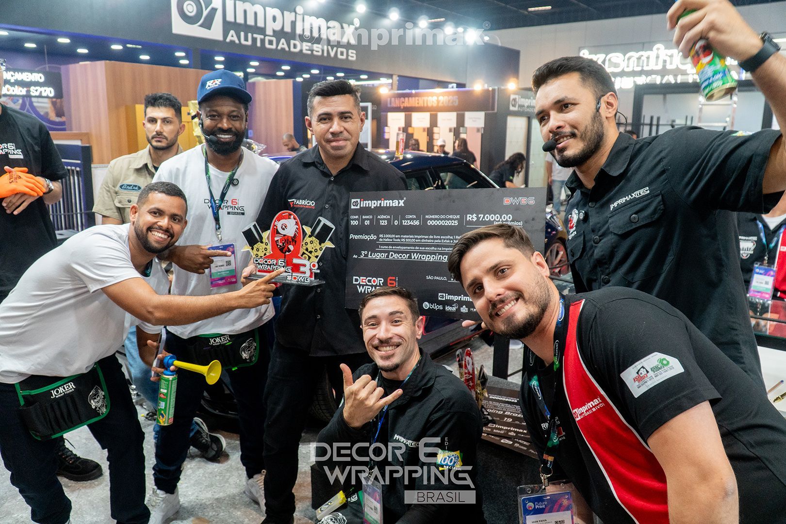 Um grupo de pessoas comemorando em um estande da Imprimax em uma feira comercial, segurando um cheque promocional e sorrindo.