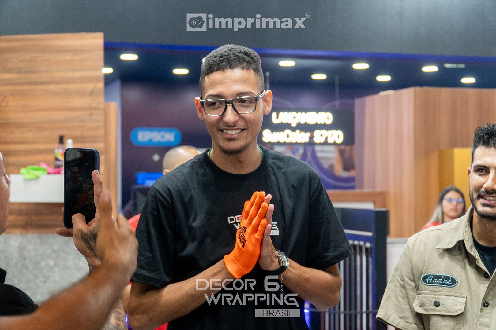 Uma pessoa sorridente, usando óculos e uma luva laranja, está em uma cabine, enquanto outra pessoa grava com um smartphone.