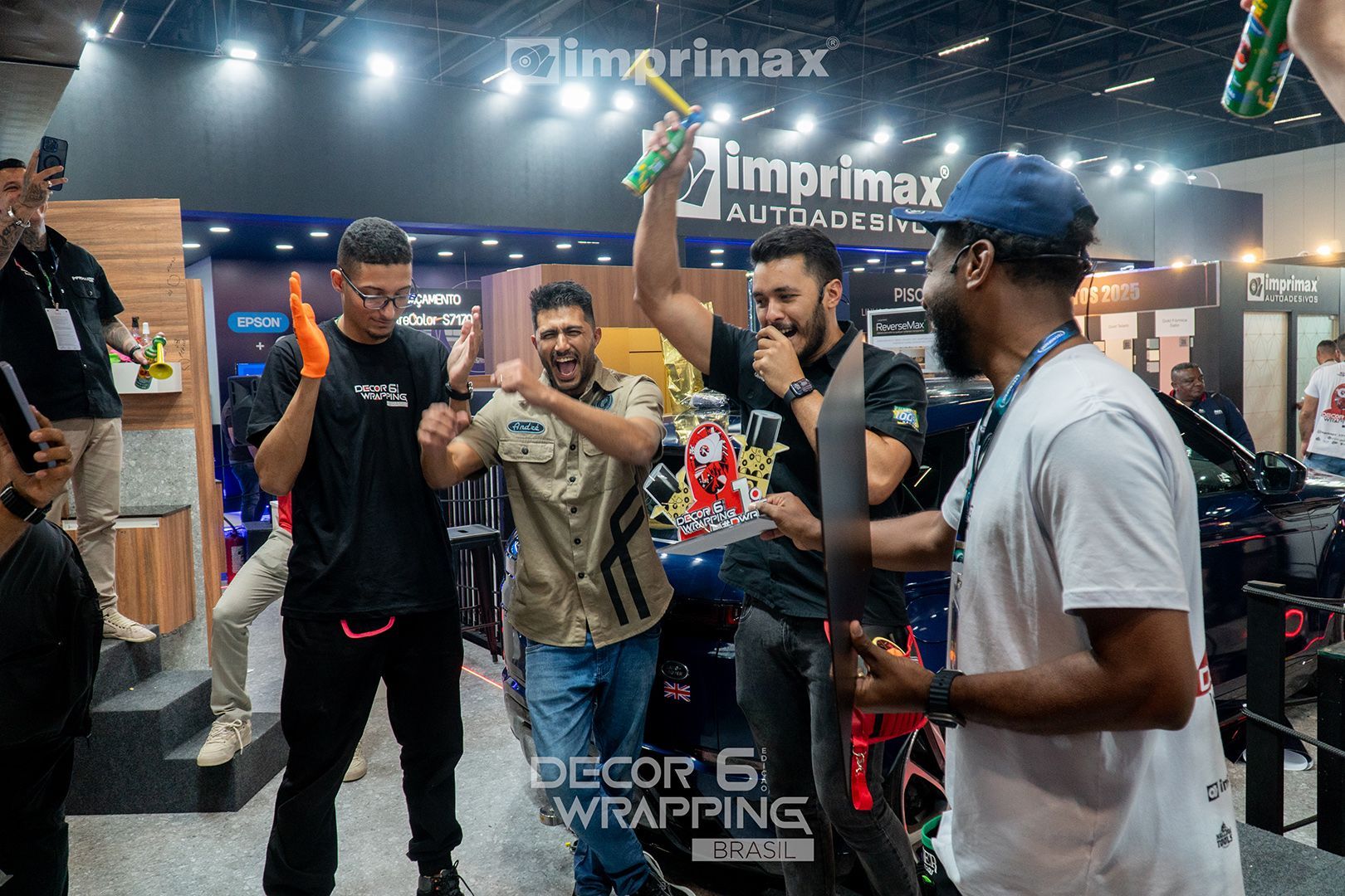 Pessoas vibrando e comemorando em um estande de feira comercial com a marca Imprimax, com uma pessoa segurando um troféu.