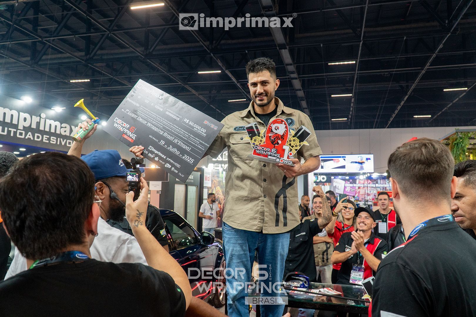 Um homem segurando um troféu e um cheque gigante em uma feira comercial da Imprimax, cercado por pessoas que o aplaudem em um salão de convenções.