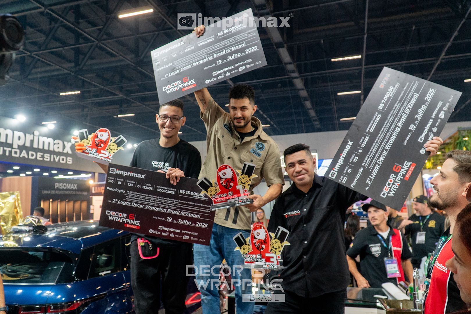 Três pessoas seguram grandes placas de premiação em um palco durante um evento em local fechado, comemorando a vitória em uma competição de envelopamento de veículos.