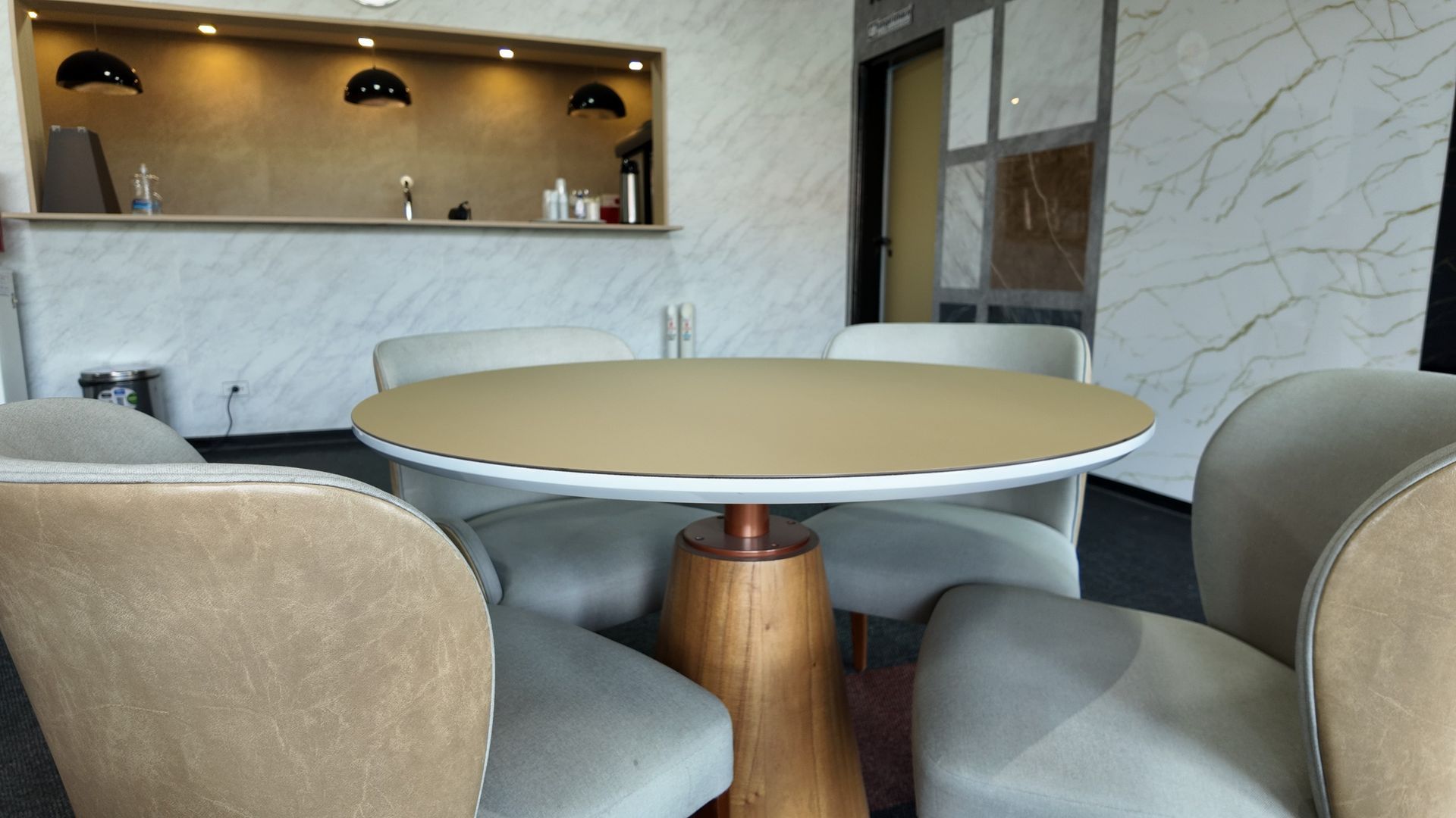 Uma mesa redonda com base de madeira rodeada por quatro cadeiras estofadas em tons claros, numa sala moderna com paredes de mármore.