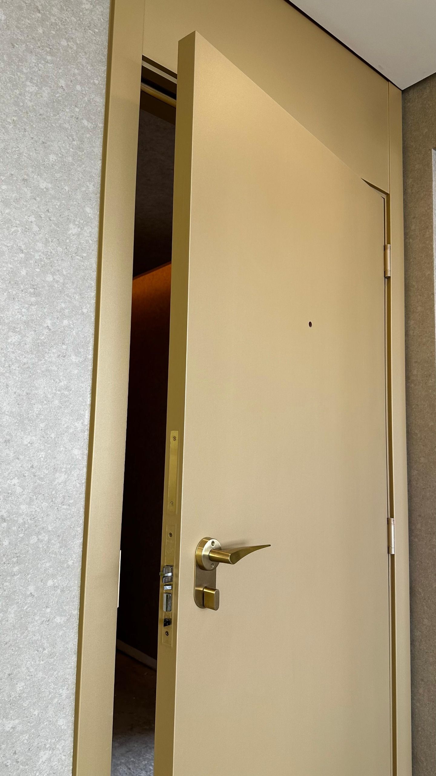 Uma porta esbranquiçada, ligeiramente entreaberta, com uma maçaneta dourada e uma olho mágico, visível contra uma parede texturizada.