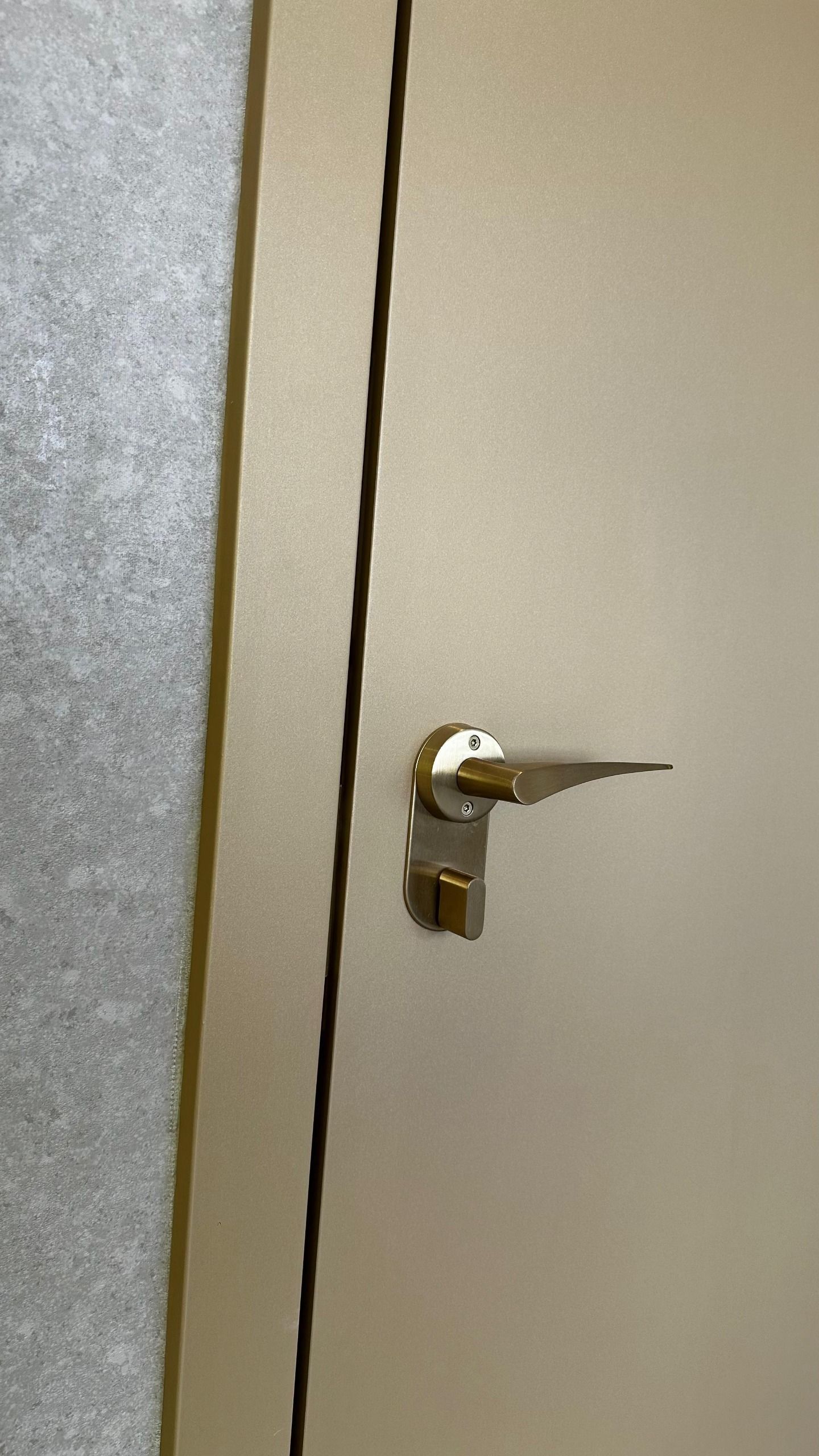 Uma visão aproximada de uma maçaneta e fechadura douradas foscas em uma porta de cor clara contra uma parede cinza texturizada.