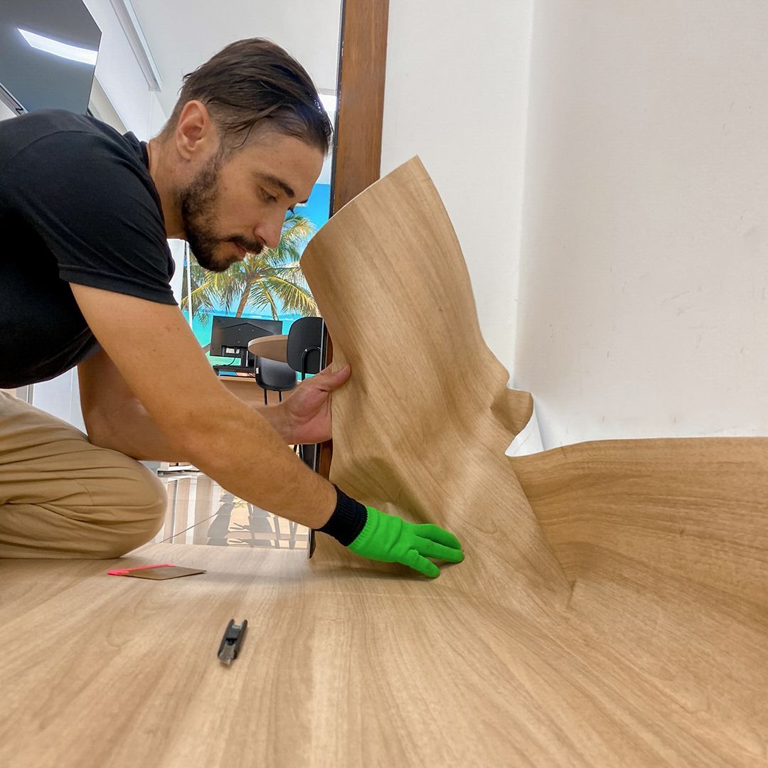 Uma pessoa vestindo camisa preta e luva verde instala piso laminado com aparência de madeira clara em um cômodo.