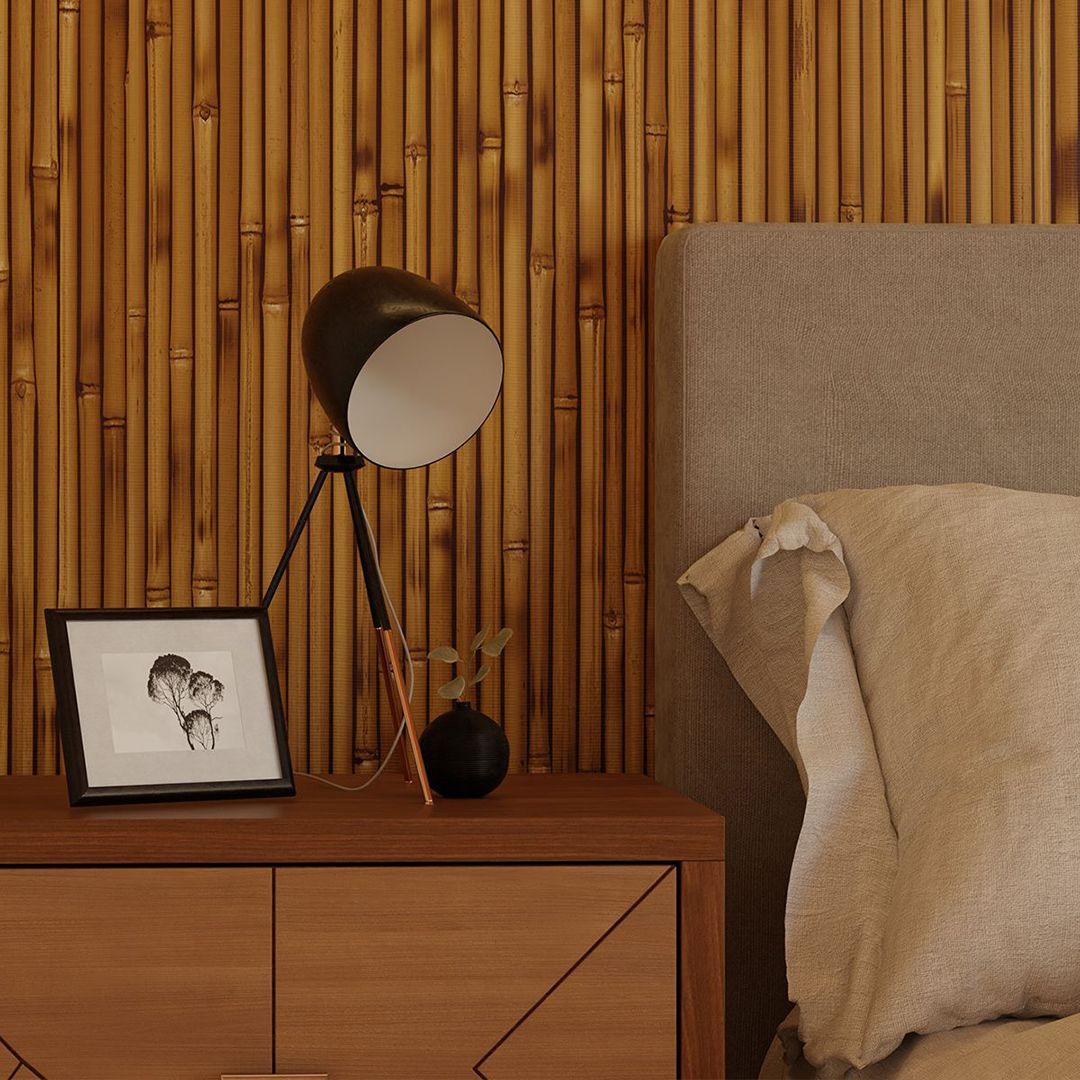 Um abajur preto, um pequeno desenho emoldurado e um vaso decorativo sobre uma mesa de cabeceira de madeira em frente a uma parede revestida de bambu.