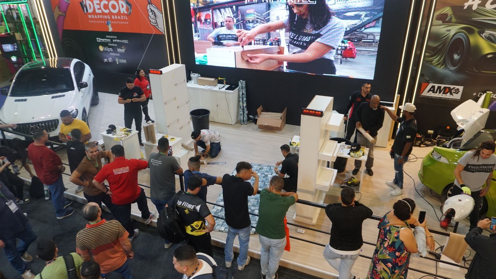 Uma multidão se reúne em torno de um estande de feira comercial que apresenta uma área de exposição, uma tela grande e pessoas examinando materiais de produtos.