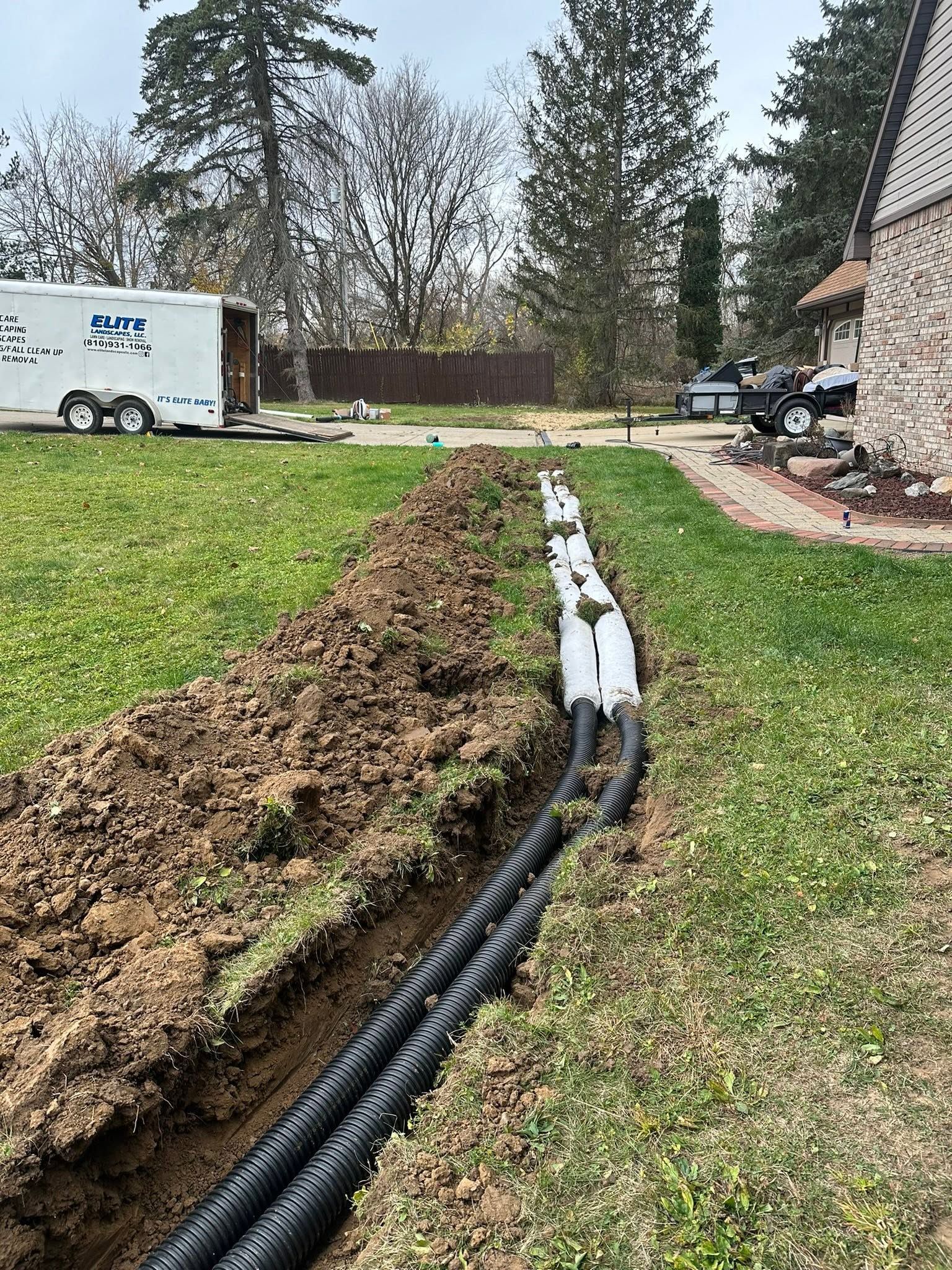 Yard with a dug trench, black drainage pipe, and a white utility trailer near a house.