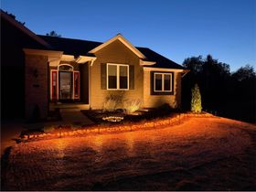 House exterior at dusk, lit by warm porch lights with orange gravel driveway and dark sky