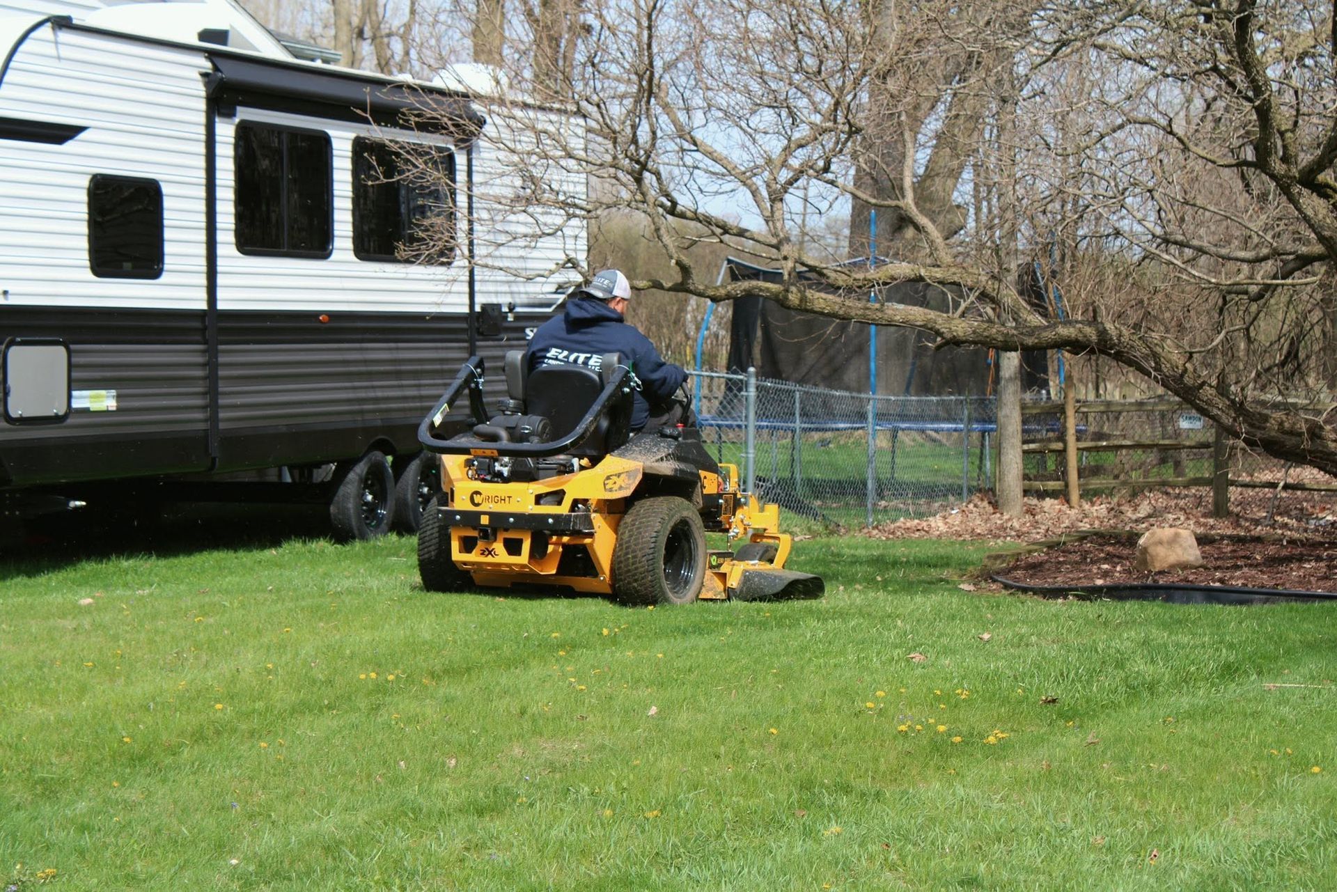 Person mowing grass beside a white RV and large tree in a yard