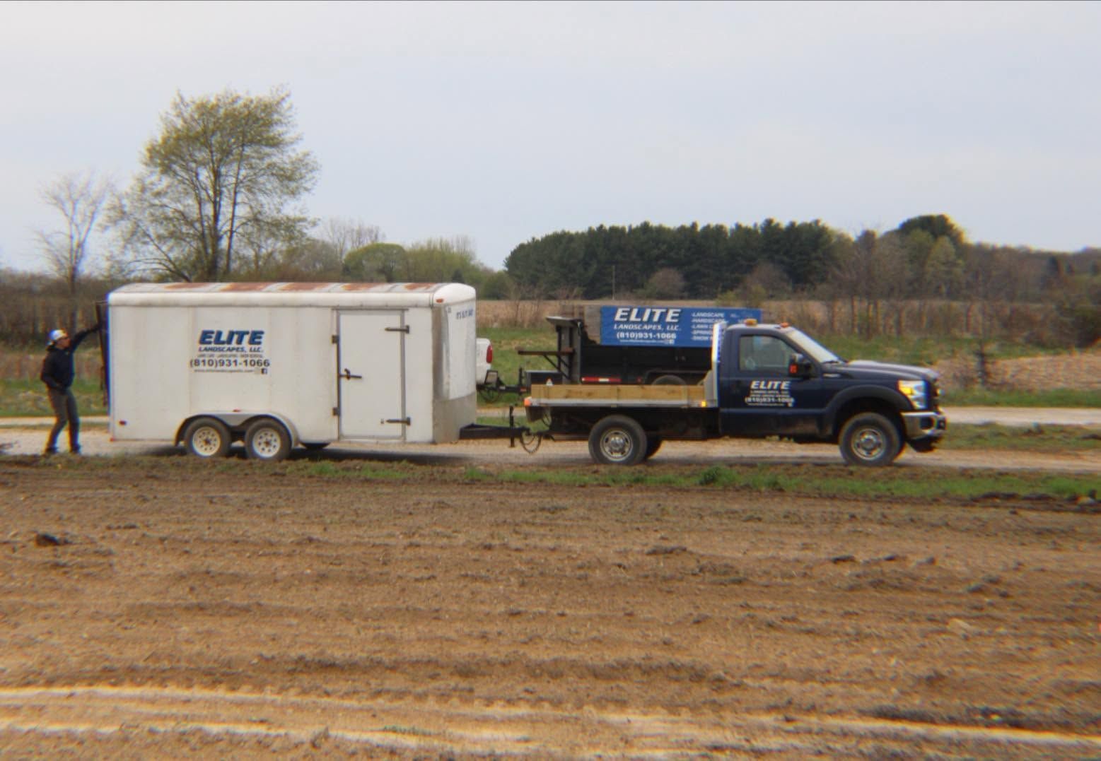 Blue pickup towing a white trailer across a muddy dirt field on a cloudy day