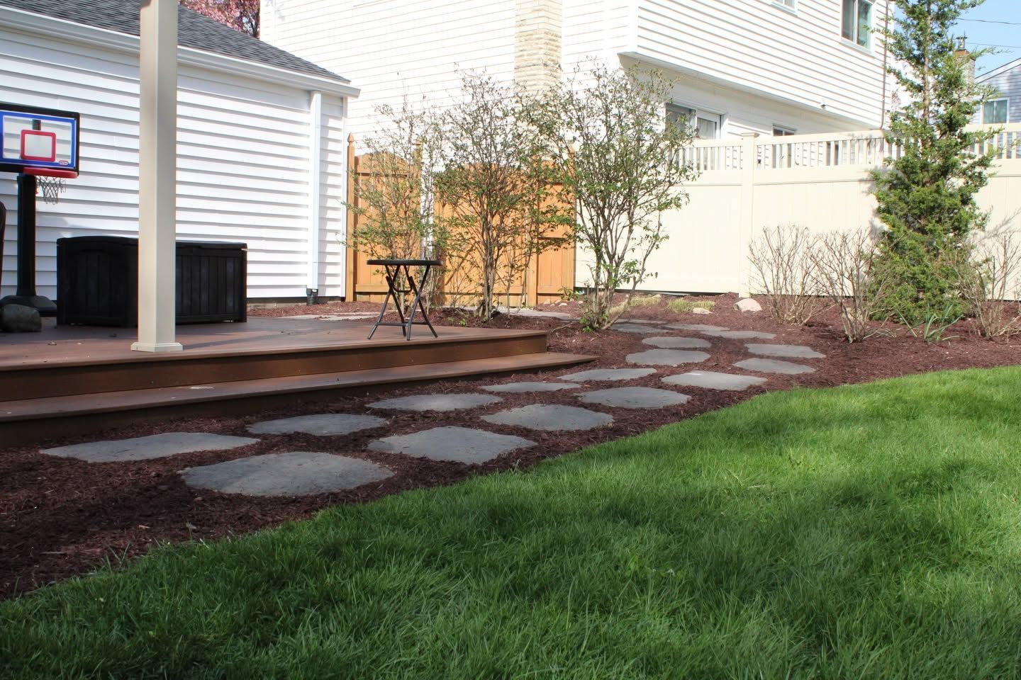 Backyard patio with stone stepping path, wooden deck, and green lawn beside a house