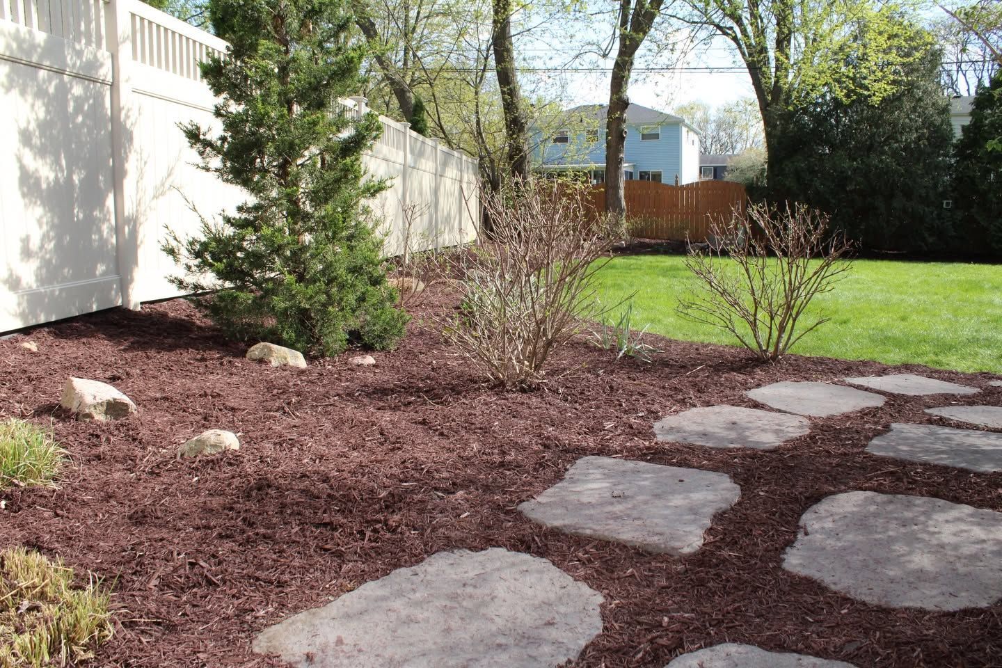 Backyard garden bed with mulch, stepping stones, shrubs, and a white fence