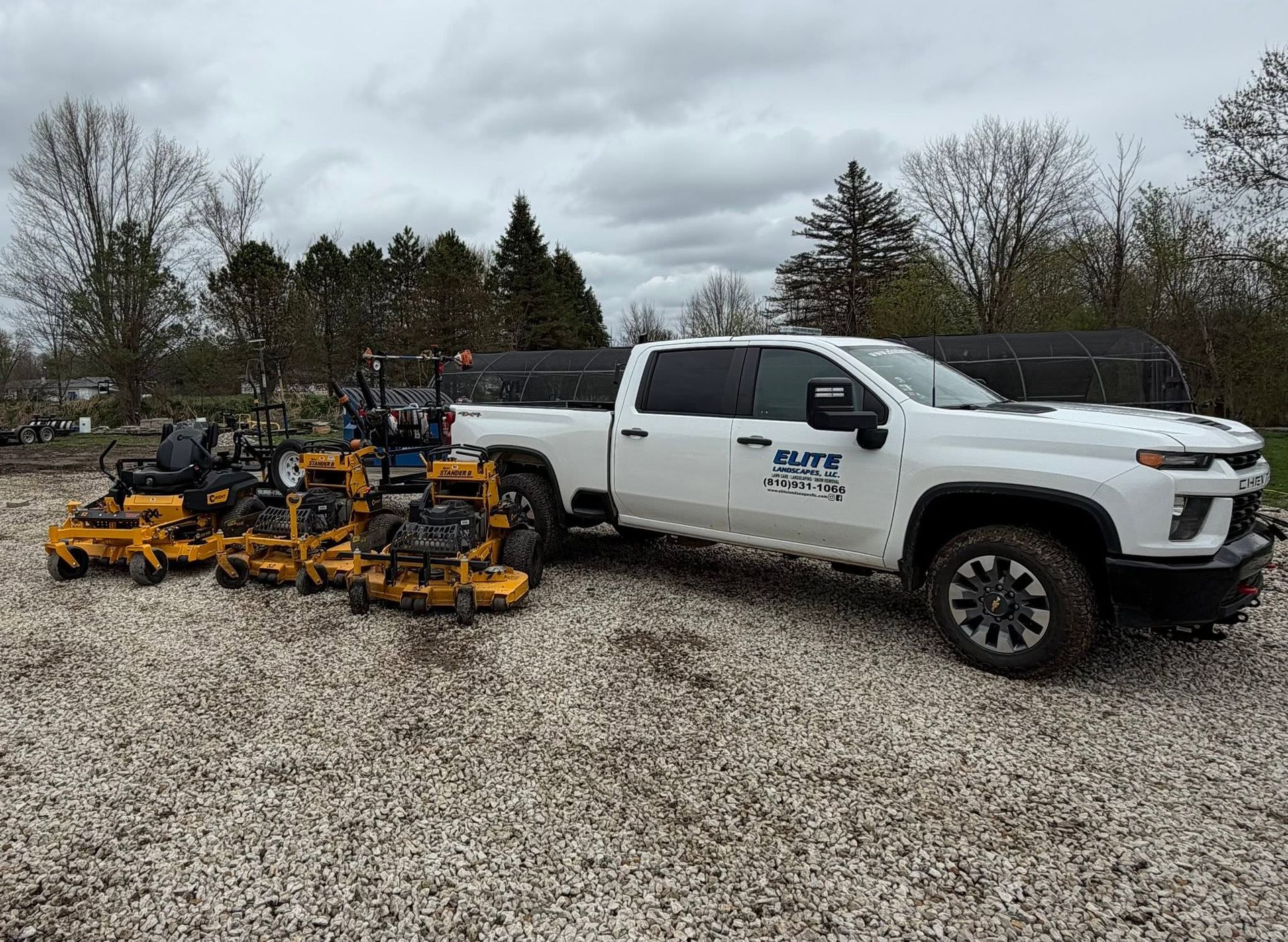White utility truck with yellow mowers parked on gravel in a leafless outdoor lot