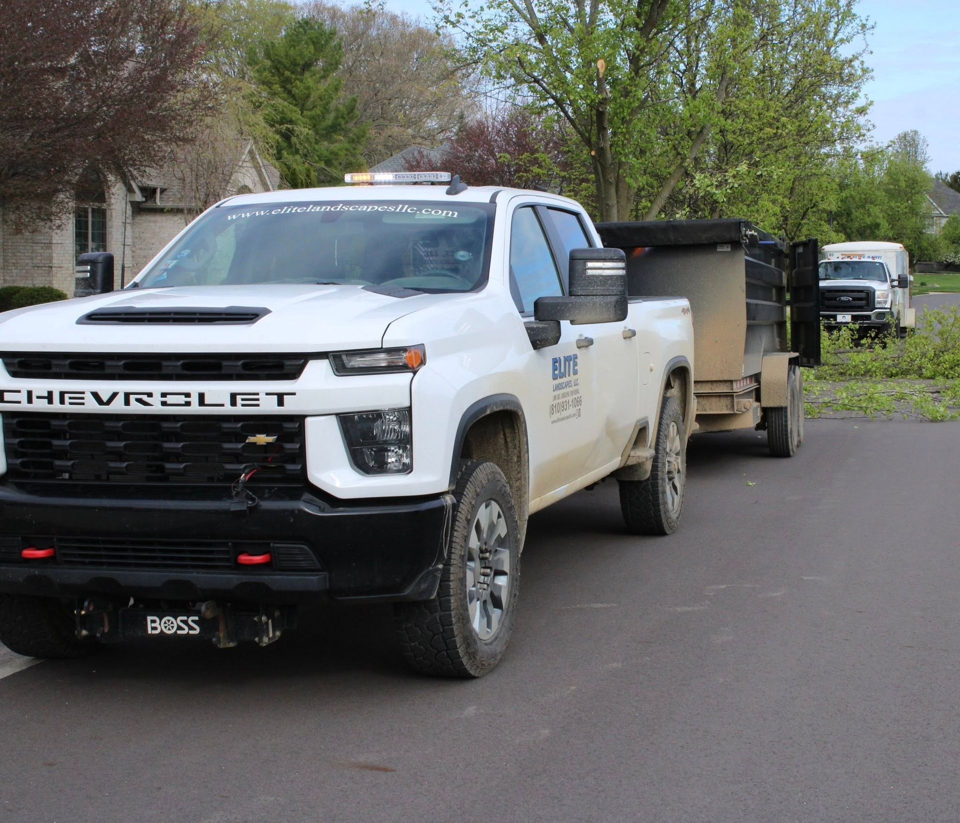 White Chevrolet pickup truck towing a black trailer on a tree-lined residential street.