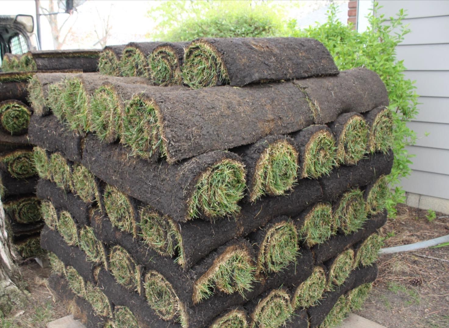Stacked rolls of sod with green grass ends, piled outdoors beside a house.
