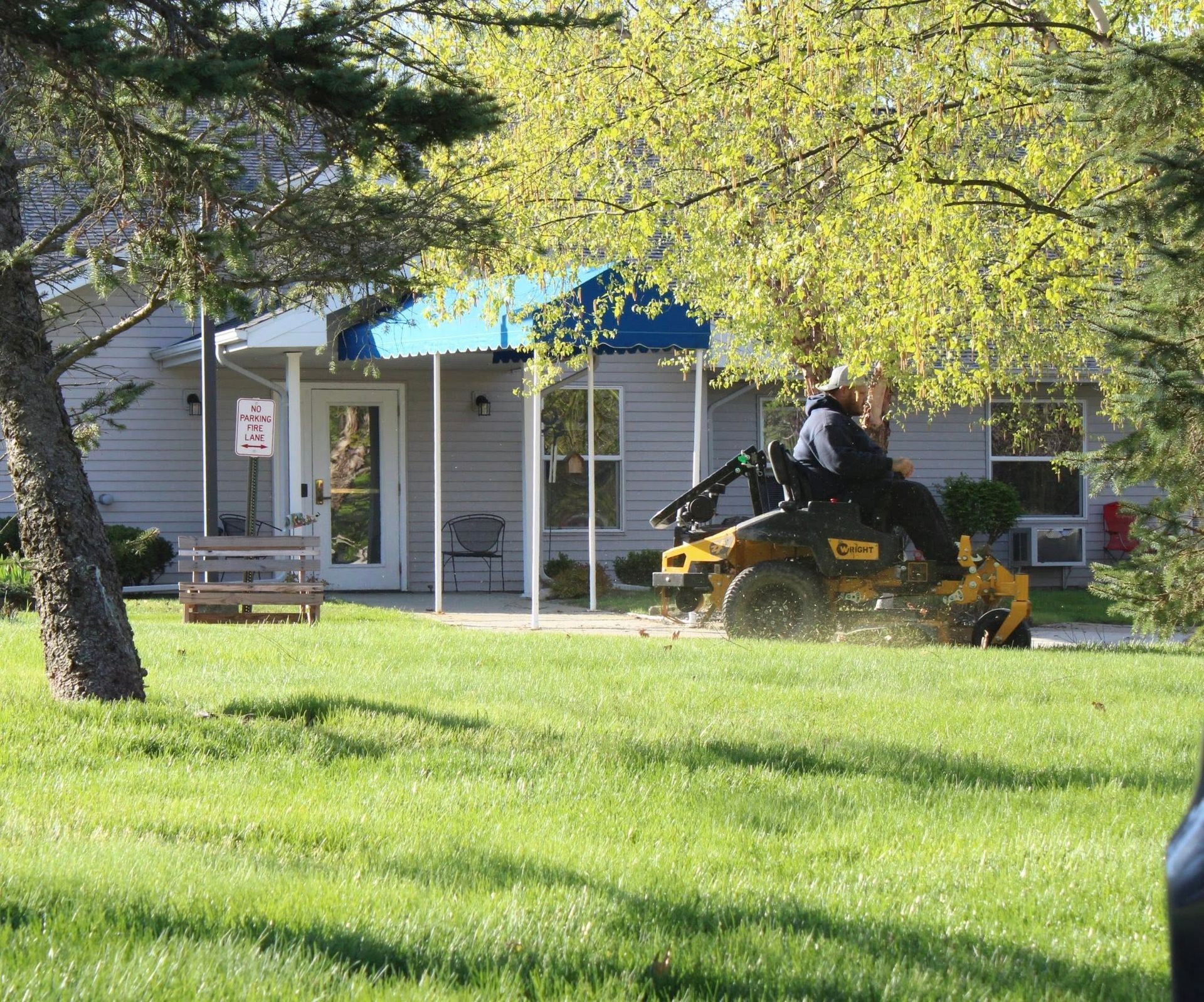Person riding a yellow lawn mower in a grassy yard beside a house under trees