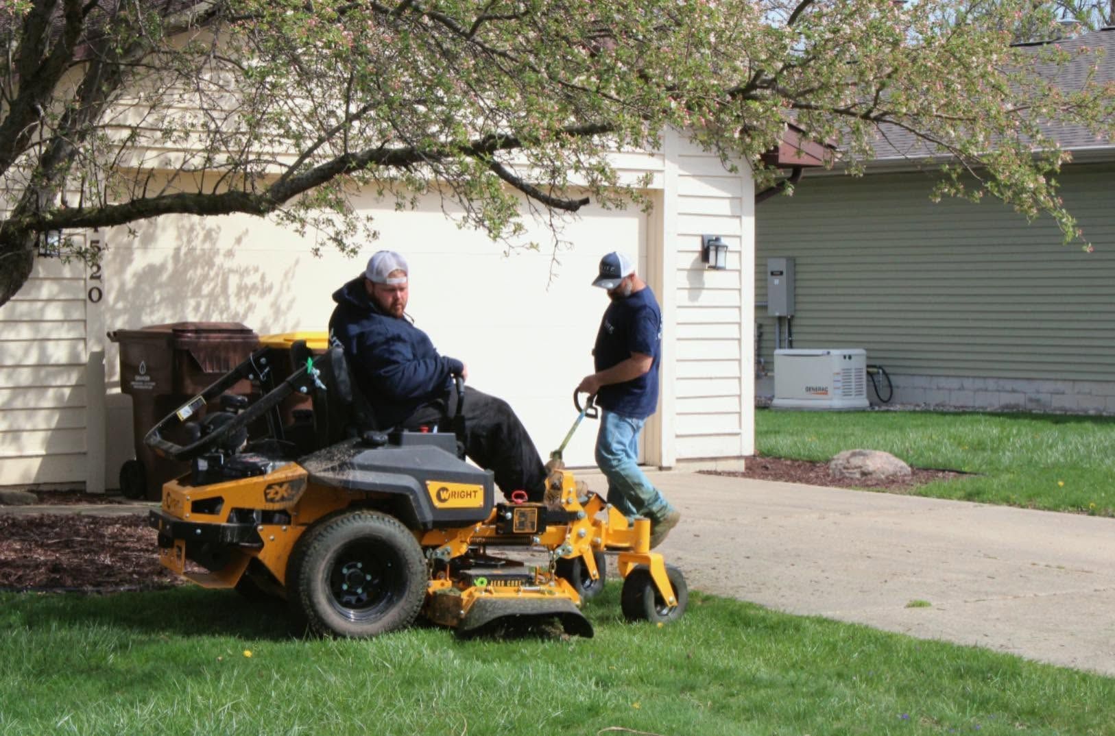 Two people on a yellow lawn mower in a suburban driveway under a tree