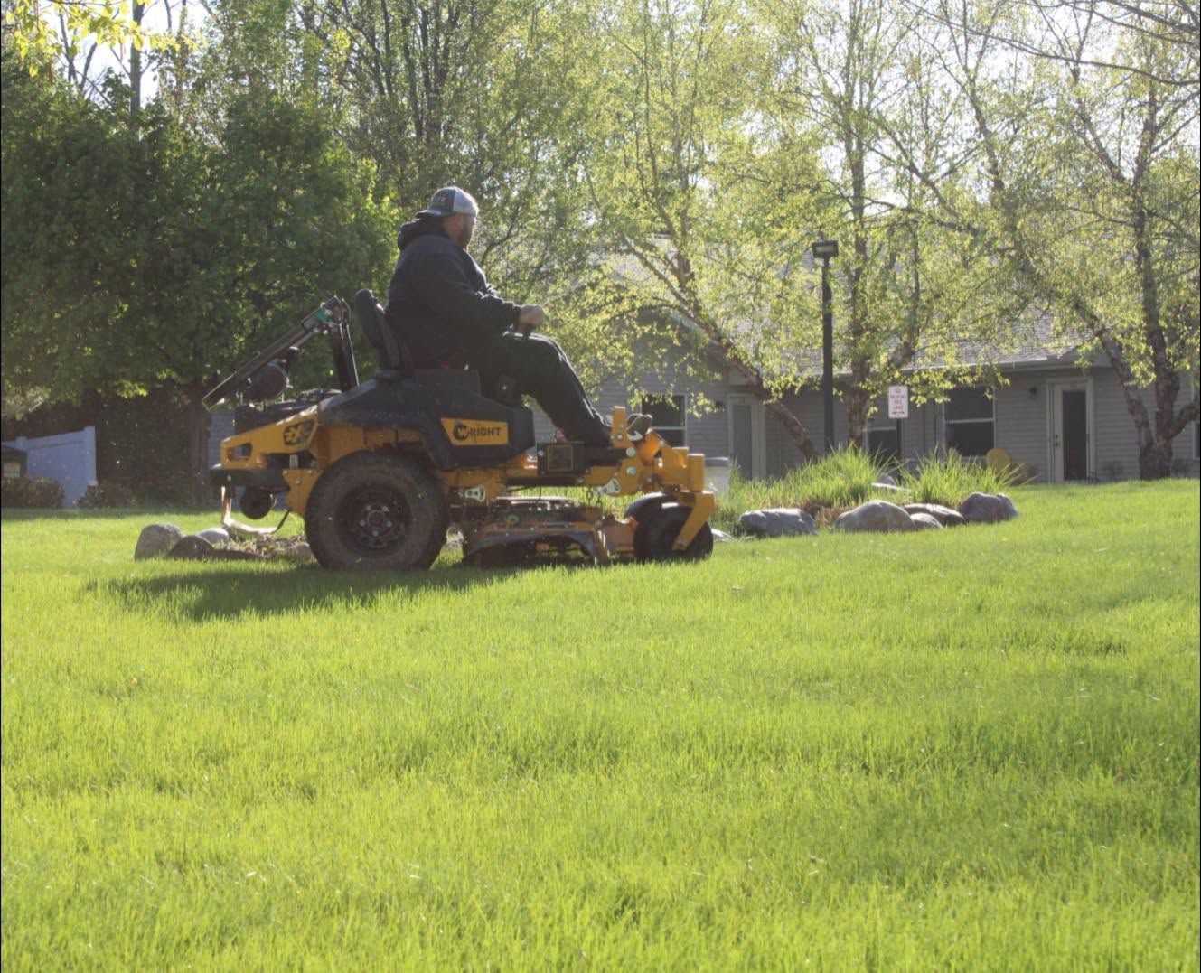 Person riding a yellow lawn mower across a green yard near a house