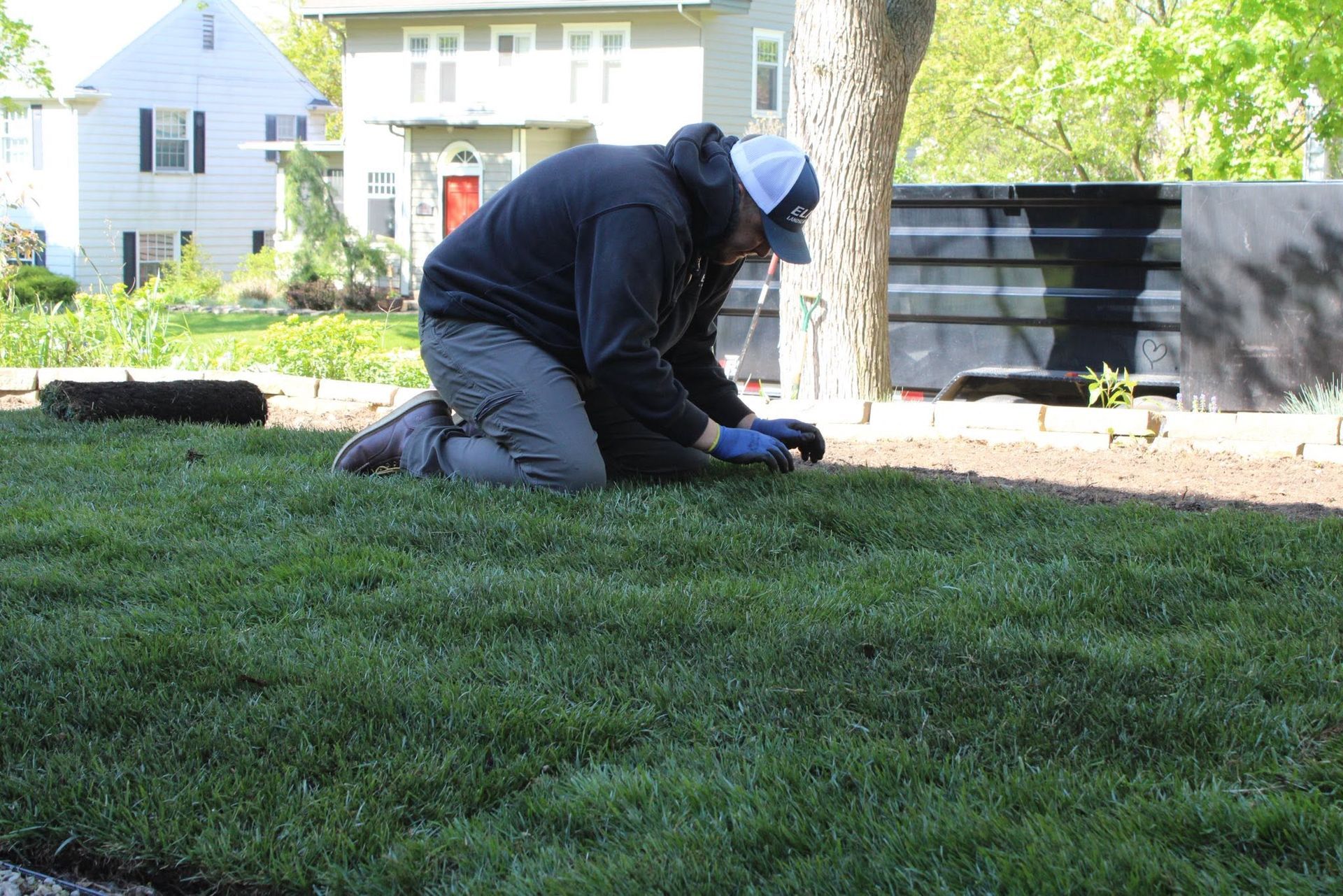 Person kneeling on grass outdoors, working near a tree and black planter boxes