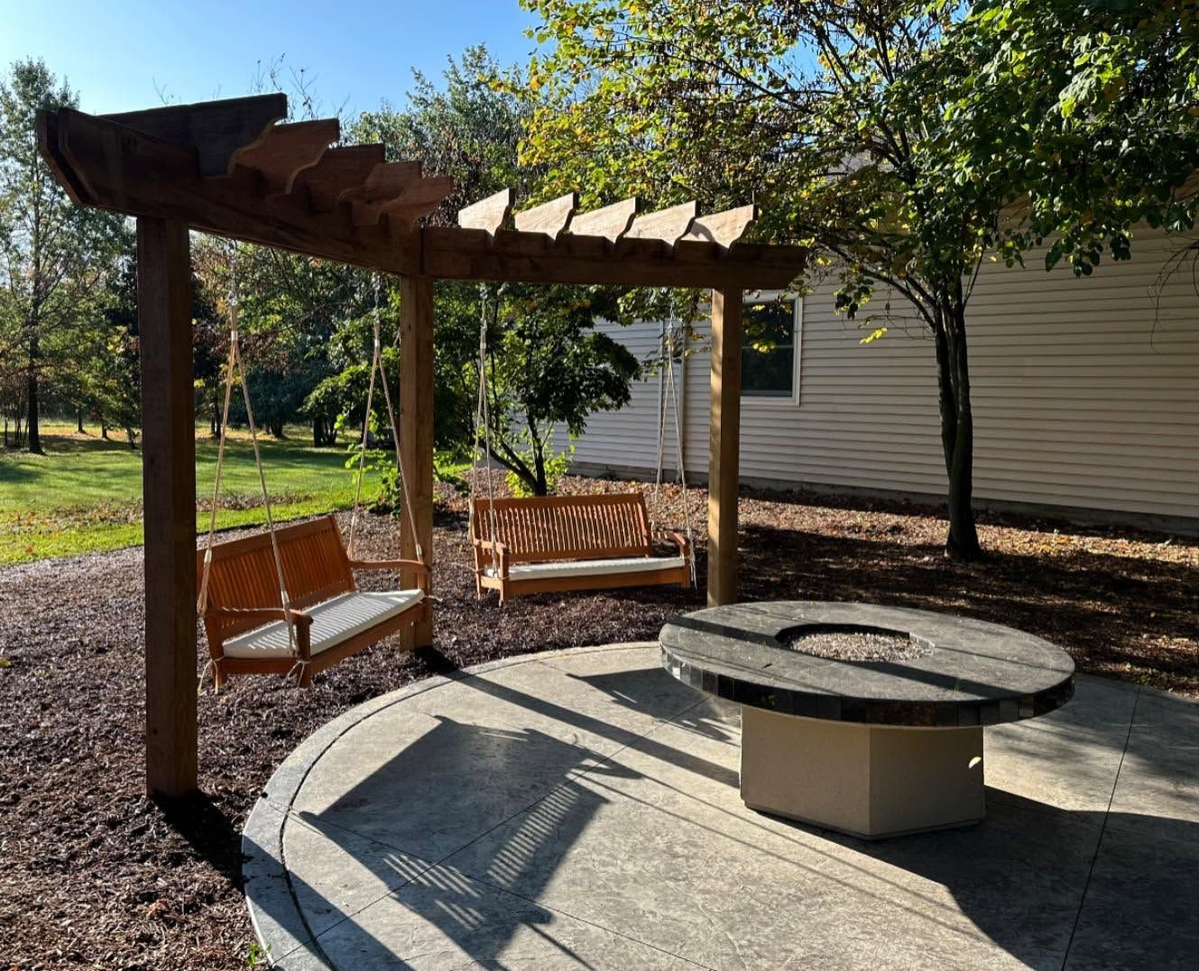 Backyard patio with pergola, wooden swings, and a circular fire pit beside a house.