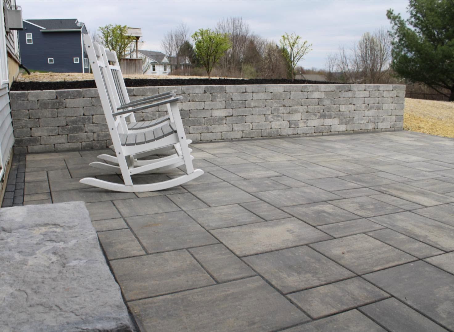White rocking chair on a gray stone patio beside a low stone wall and open yard.