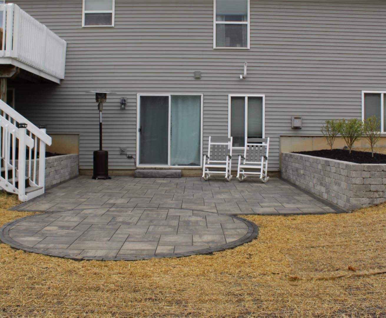 Stone patio with white rocking chairs against a house, beside a patchy lawn and small retaining wall.
