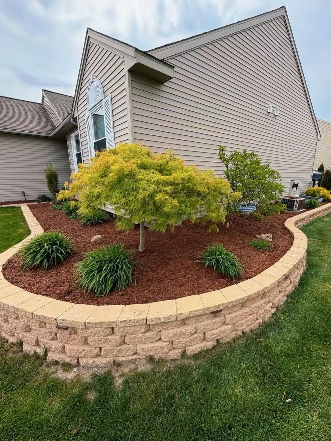 Landscaped front yard with a small tree, shrubs, mulch, and a curved stone retaining wall beside a house
